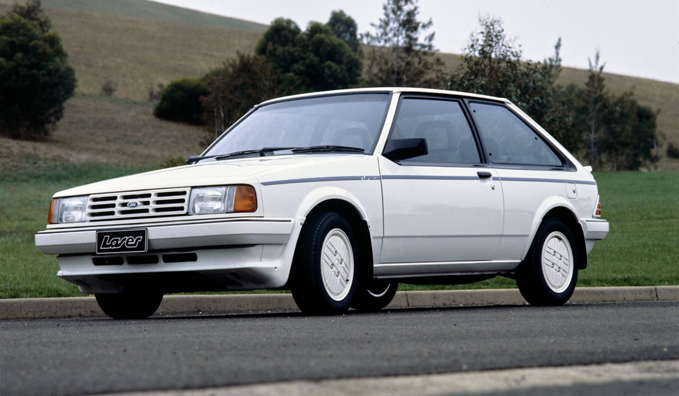 Front three-quarter view of a white 1980s Ford Laser 2‑door hatchback parked on a paved road with a grassy field behind it in the countryside.