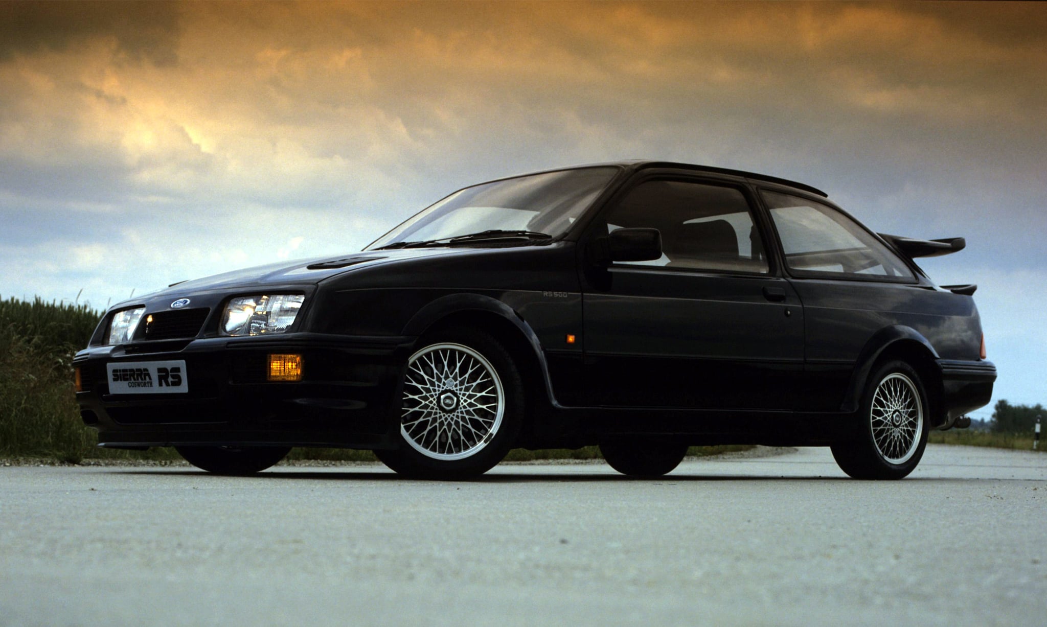 Black Ford Sierra RS Cosworth hatchback parked on a road under a dramatic cloudy sky, front three-quarter view with distinctive mesh wheels.