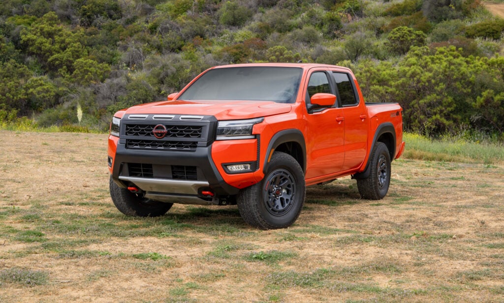 Orange pickup truck parked on a dirt field with green hills in the background, front three-quarters view showing rugged tires and grille rail.