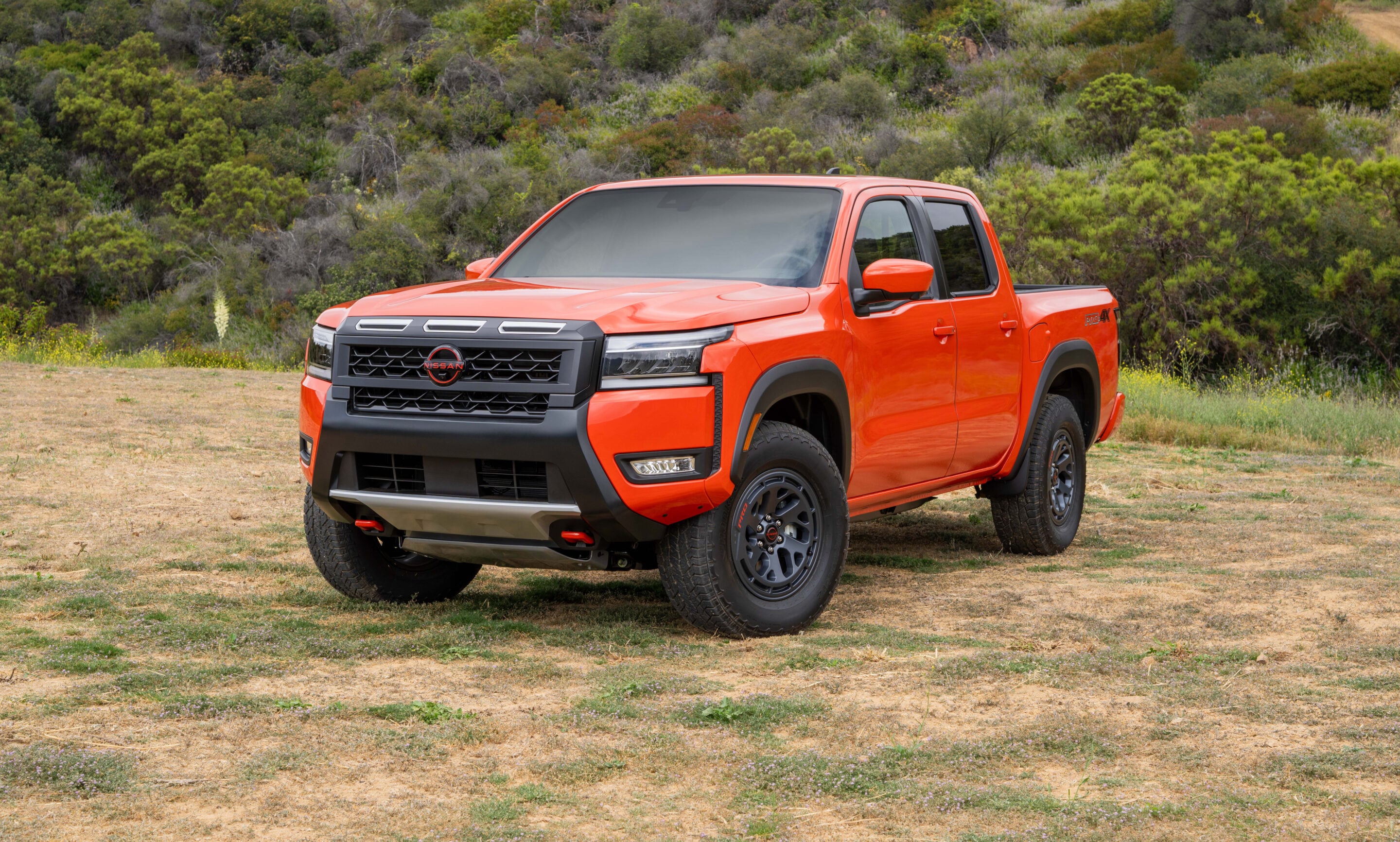 Orange pickup truck parked on a dirt field with green hills in the background, front three-quarters view showing rugged tires and grille rail.