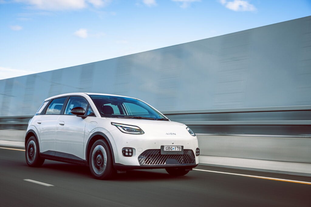 White electric SUV (AION) driving on a highway beside a concrete barrier against a blue sky with light clouds, motion blurred background.