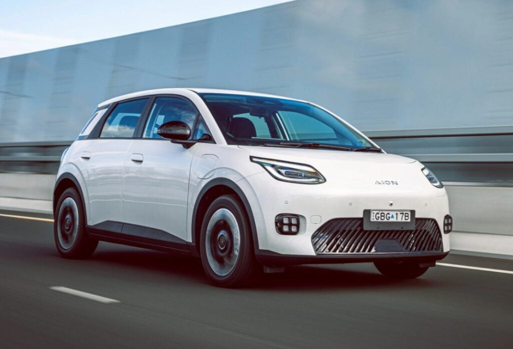 White compact electric car driving on a highway, front grille with distinctive lower venting and sleek headlights, mid-mand speed blur in background.