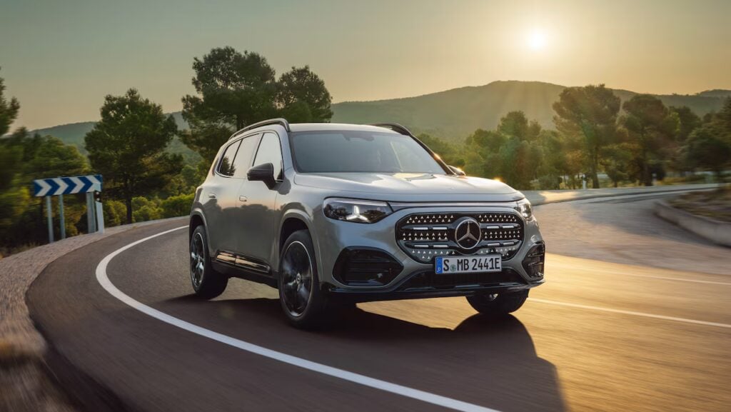 Silver Mercedes-Benz SUV driving on a winding mountain road at sunset, trees in the background and warm light reflection on the car surface.