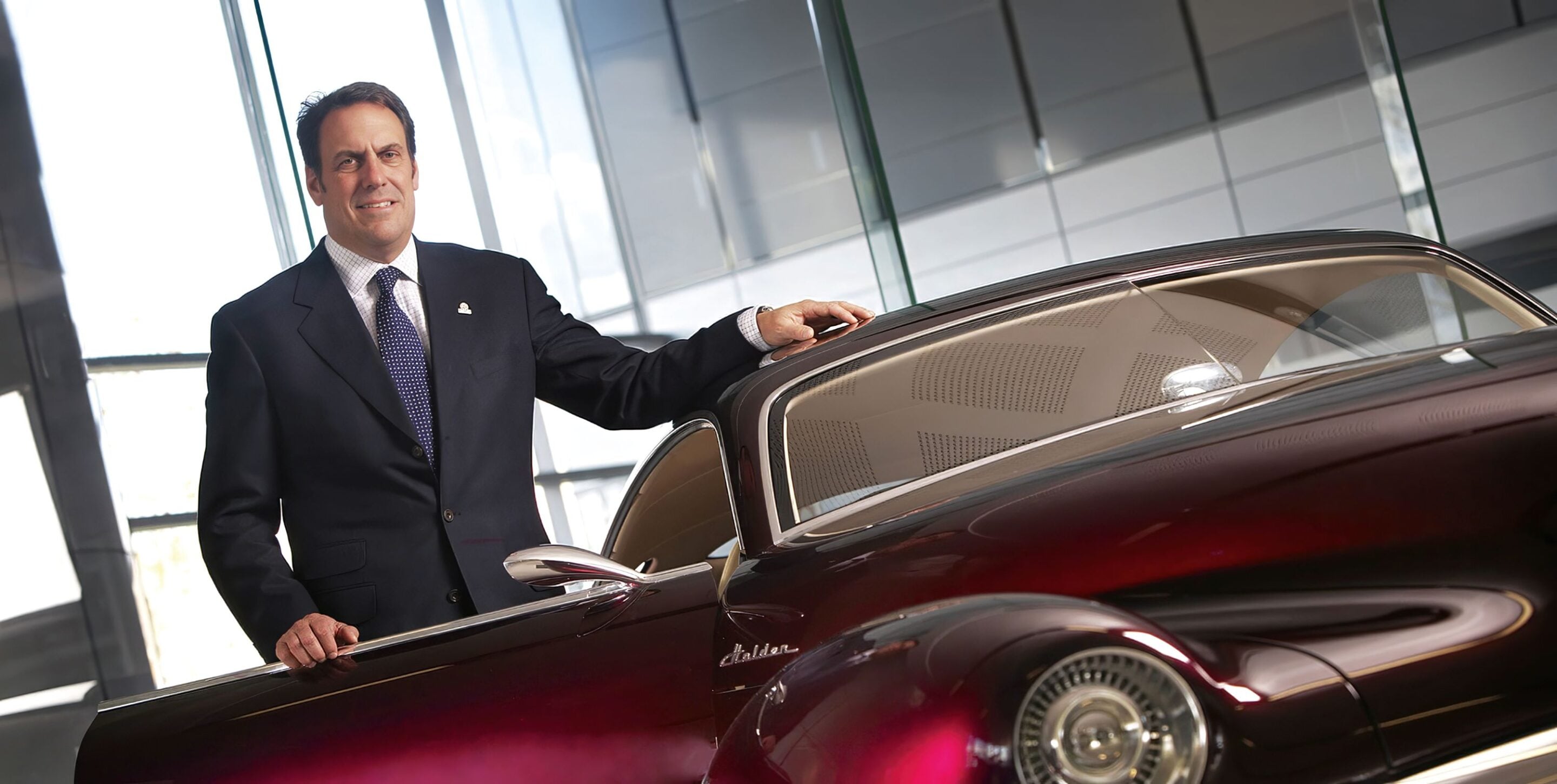 Businessman in a navy suit leans on a burgundy vintage car inside a showroom.