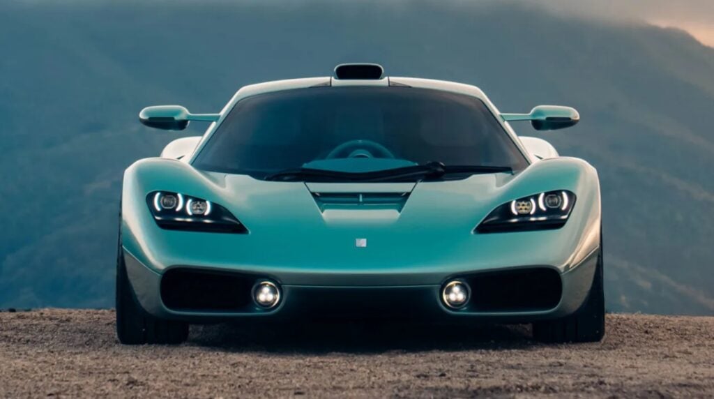Front view of a turquoise sports car on a dirt road with mountains in the background, headlights on.