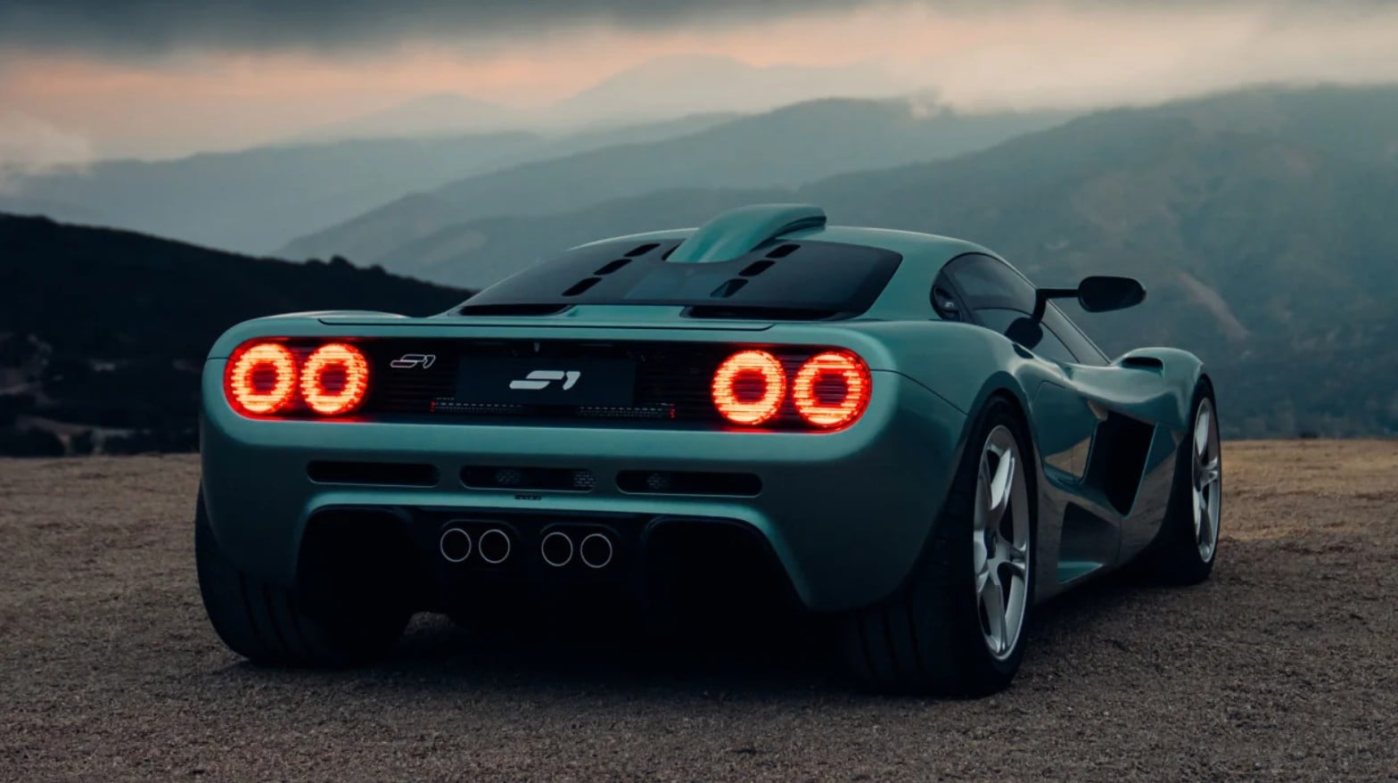 Blue mid-engine sports car seen from rear, parked on a dusty hill with mountains in the background, taillights glowing red.