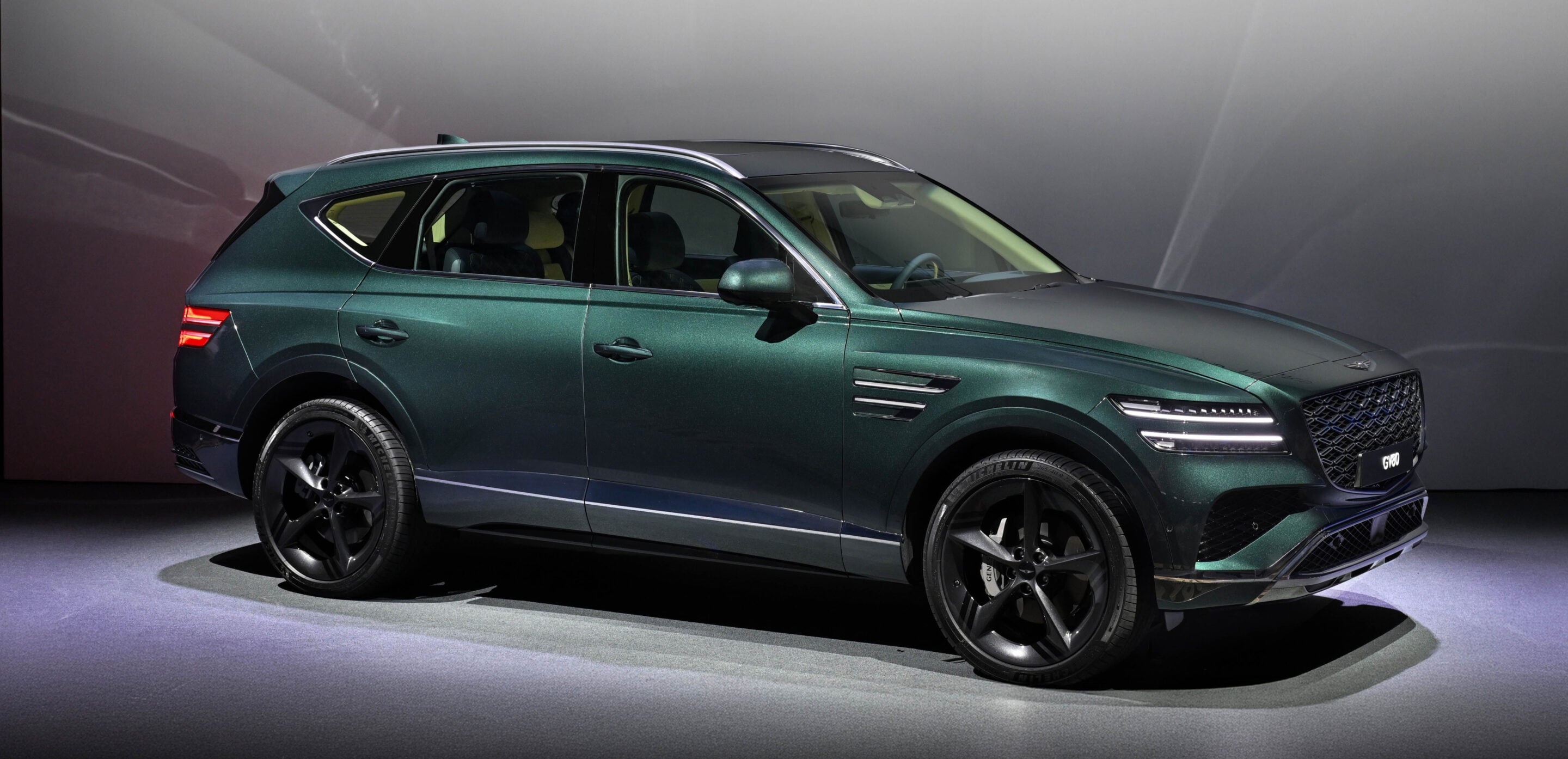 Green Genesis GV80 SUV on a showroom floor, angled side view with sunroof visible.