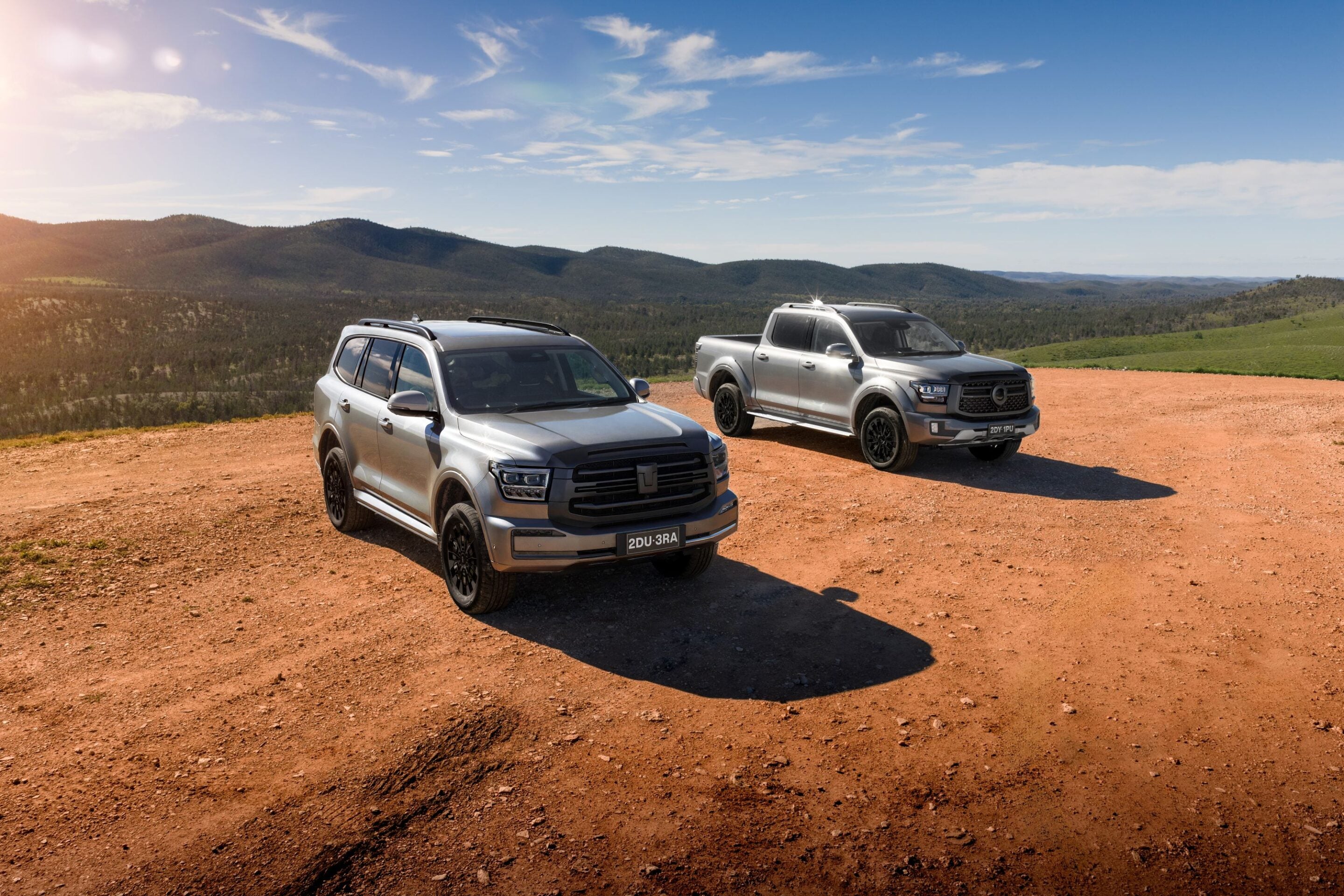 Two silver pickup trucks parked on a red dirt road against a backdrop of rolling hills and a blue sky.