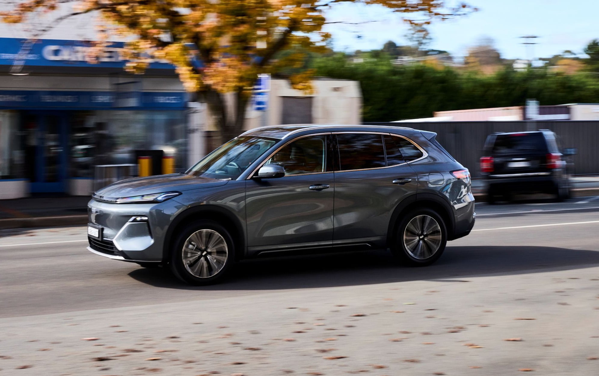 Gray SUV driving down a city street with blurred storefronts in the background, autumn leaves on the road indicating movement and speed.