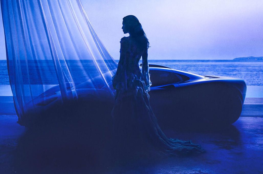Silhouetted woman in an elegant gown stands beside a sleek car by the ocean, bathed in blue light at dusk.