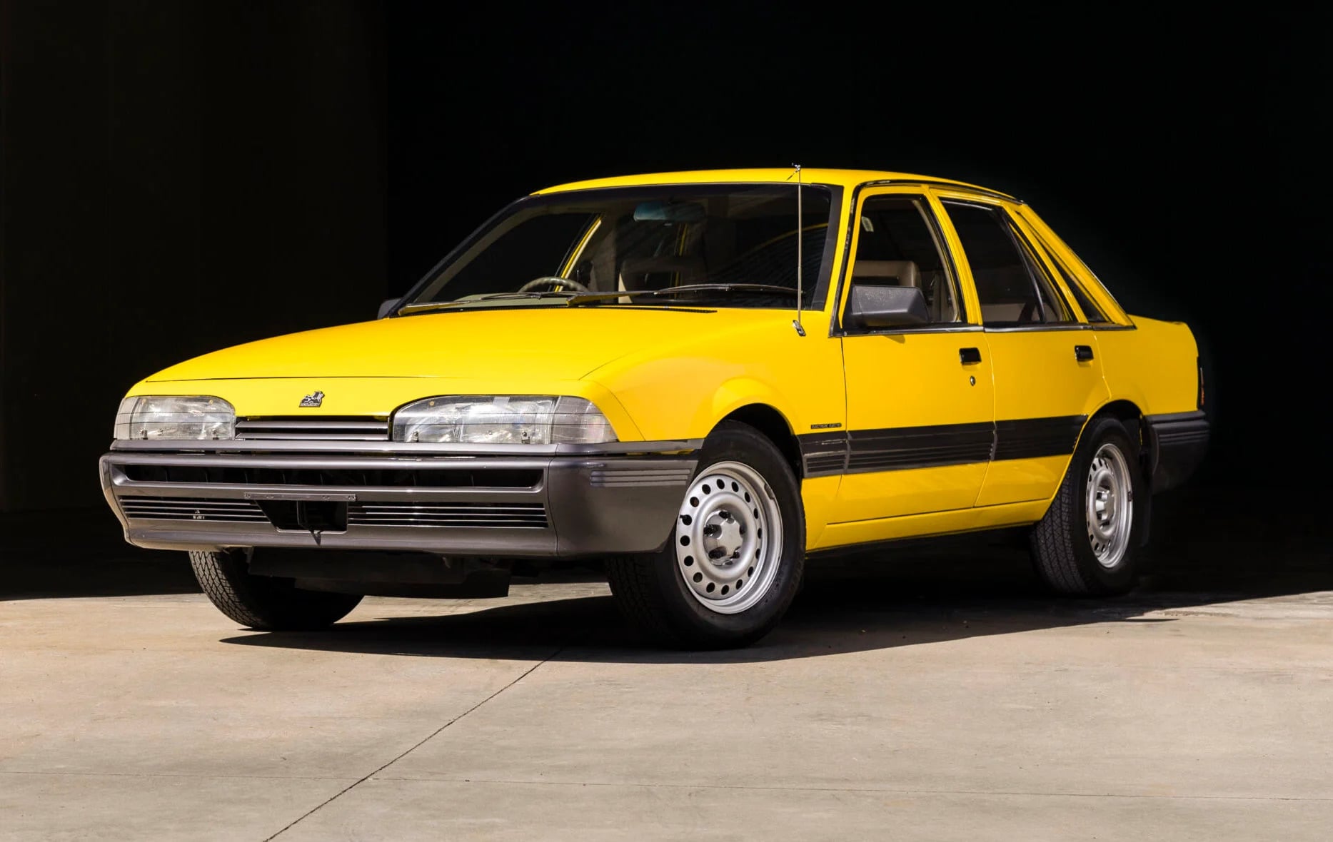 Yellow vintage four-door station wagon with black trim, parked on a light concrete surface against a dark background.
