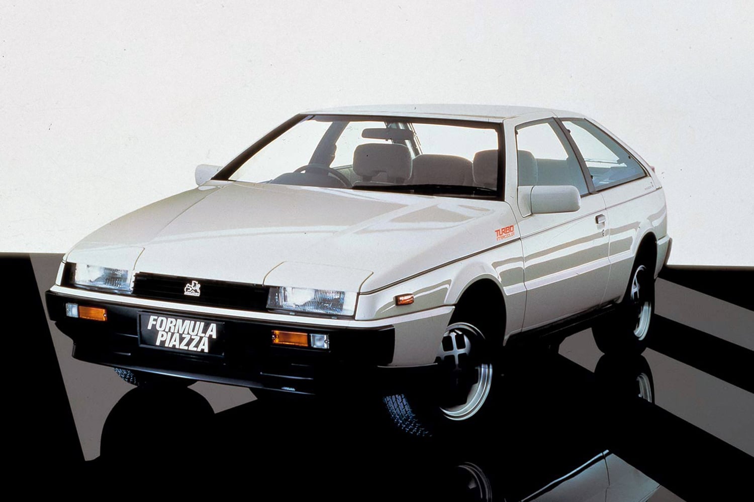 White compact sports coupe shown in a three-quarter front view, with a 'FORMULA PIAZZA' license plate, photographed in a studio.
