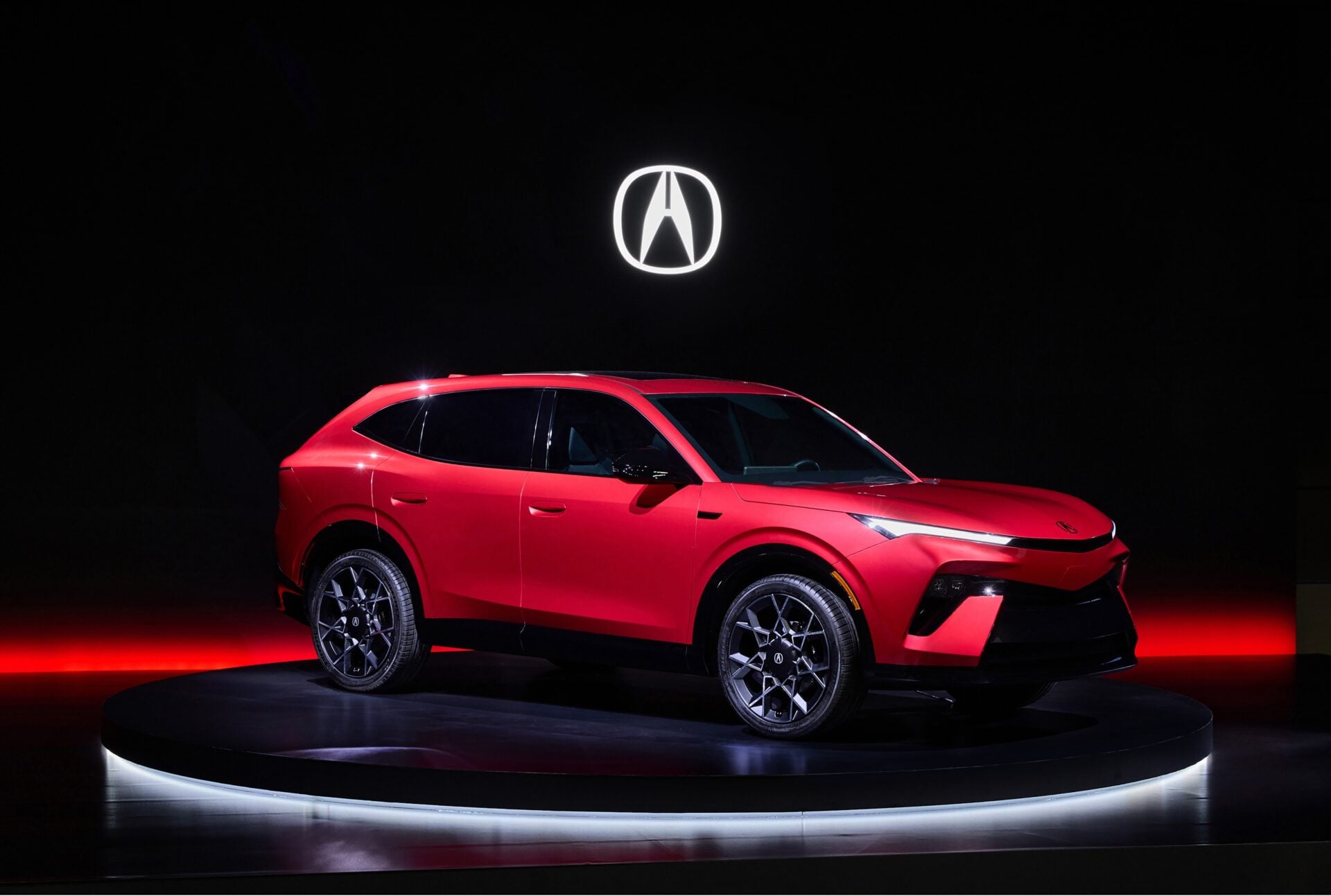 Red SUV on a circular display platform with a glowing Acura logo above in a dark showroom setting.