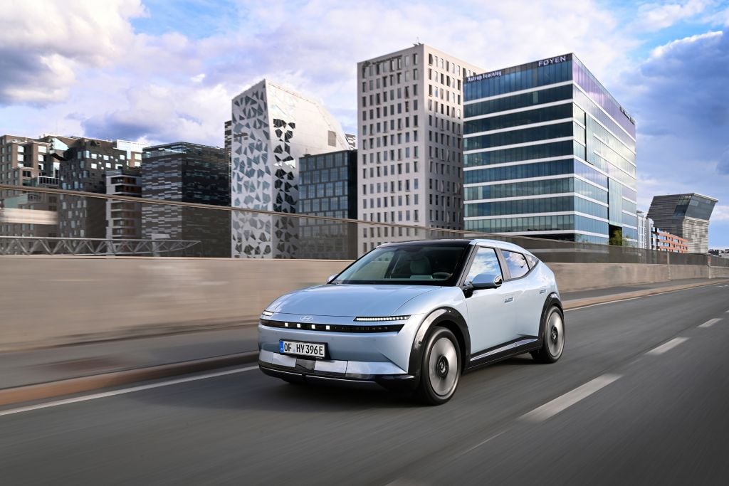 Silver electric SUV driving on a city highway with modern glass buildings in the background under a blue sky.