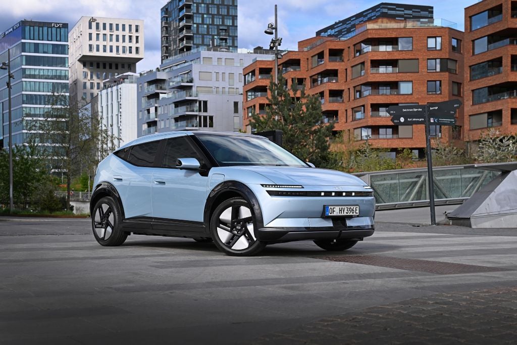Light blue electric SUV parked on a paved plaza with modern buildings in the background.
