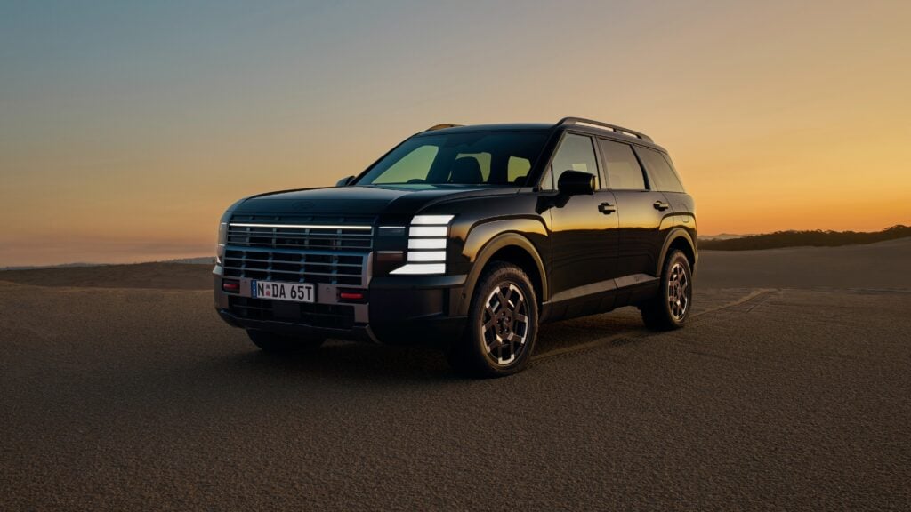 Black SUV parked on a desert road at sunset, warm orange sky and distant dunes behind it.