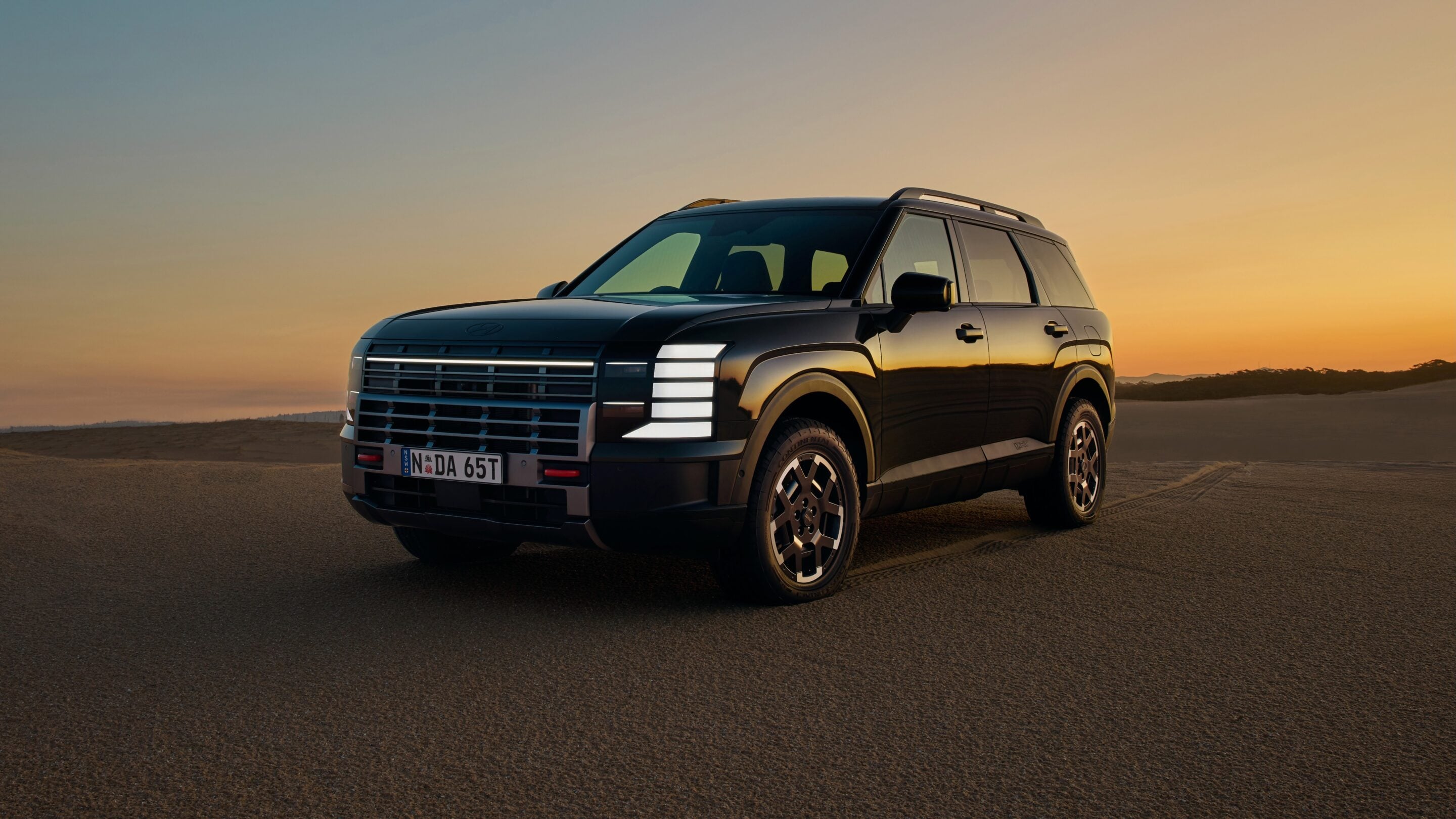 Black SUV parked on a desert road at sunset, warm orange sky and distant dunes behind it.