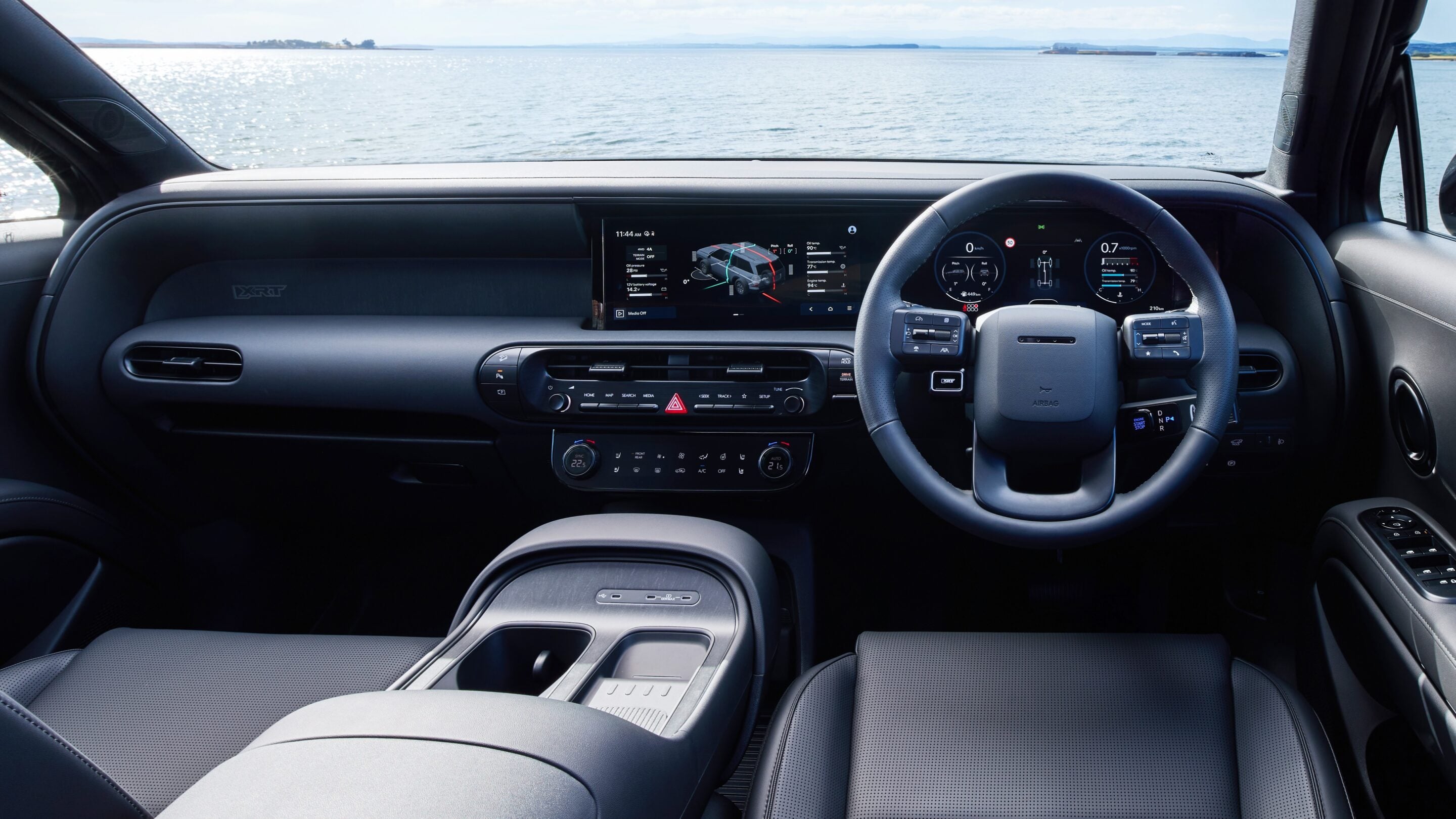 Inside a car cockpit with a large central touchscreen, digital gauges on the instrument cluster, and ocean scenery visible through the windshield.
