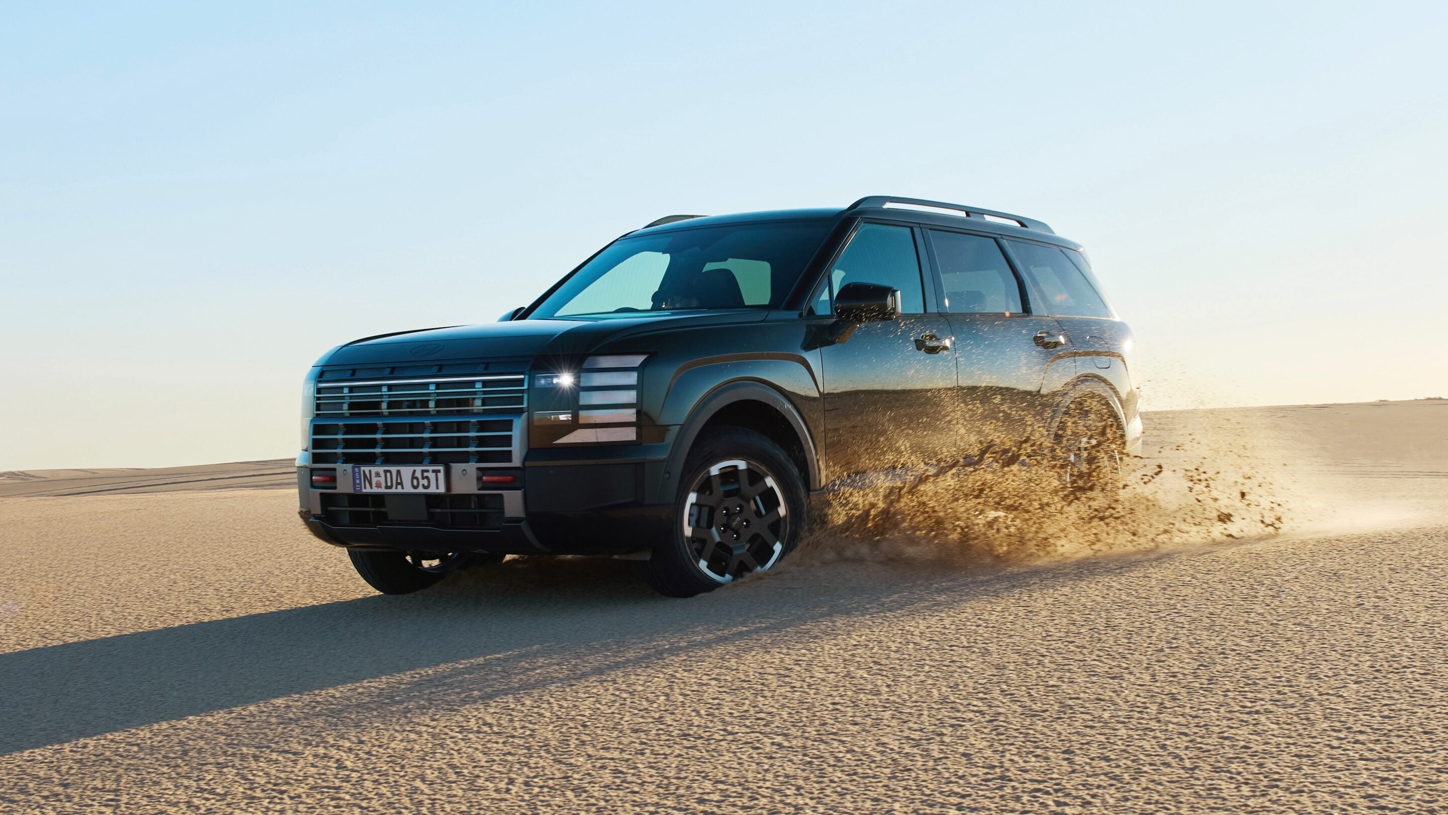 Black SUV speeding across a sandy desert, sand spraying from the tires under a clear blue sky.