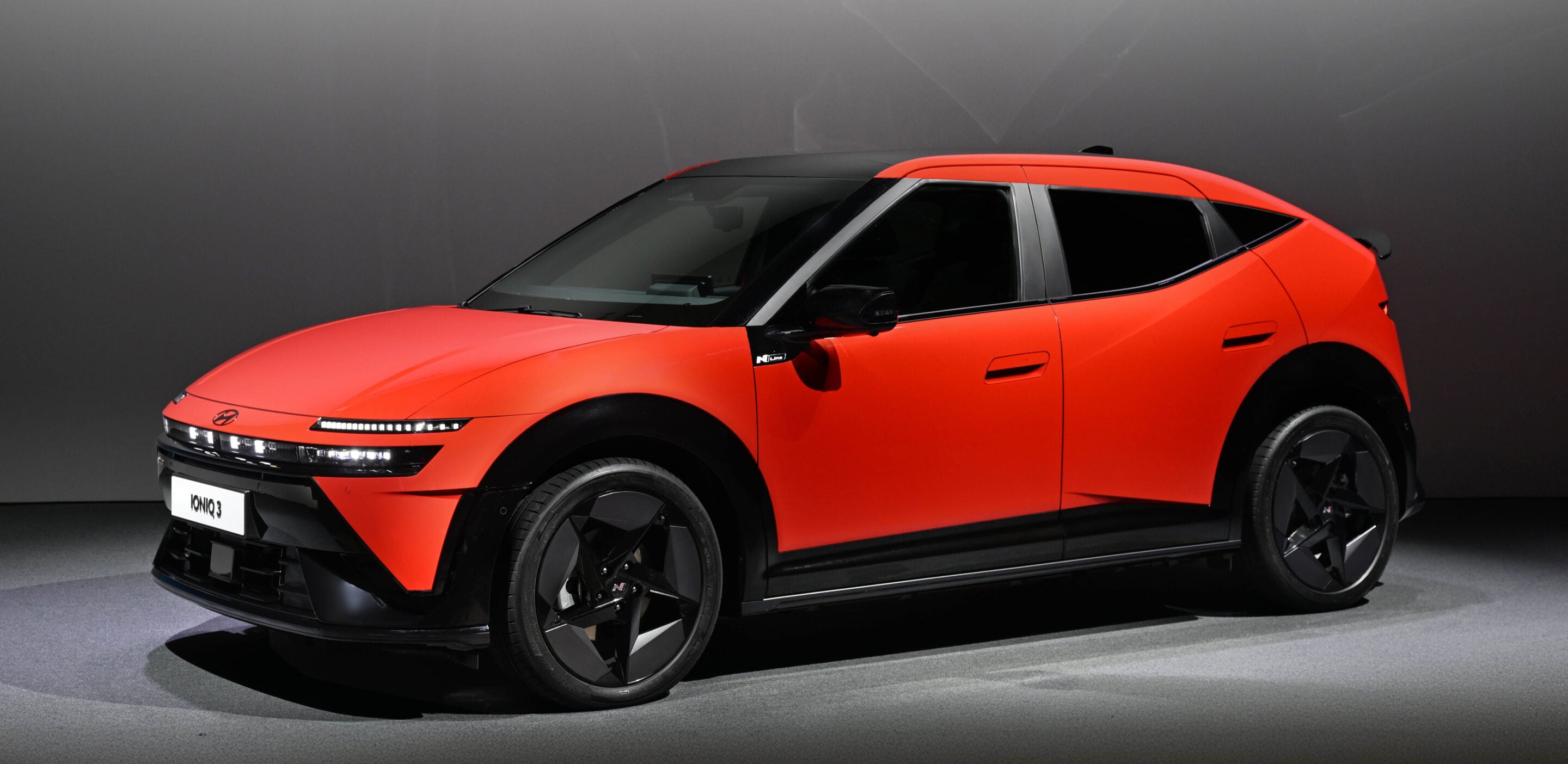 Orange electric SUV on display in a studio, shown from a front-left angle with black trim and large wheels.