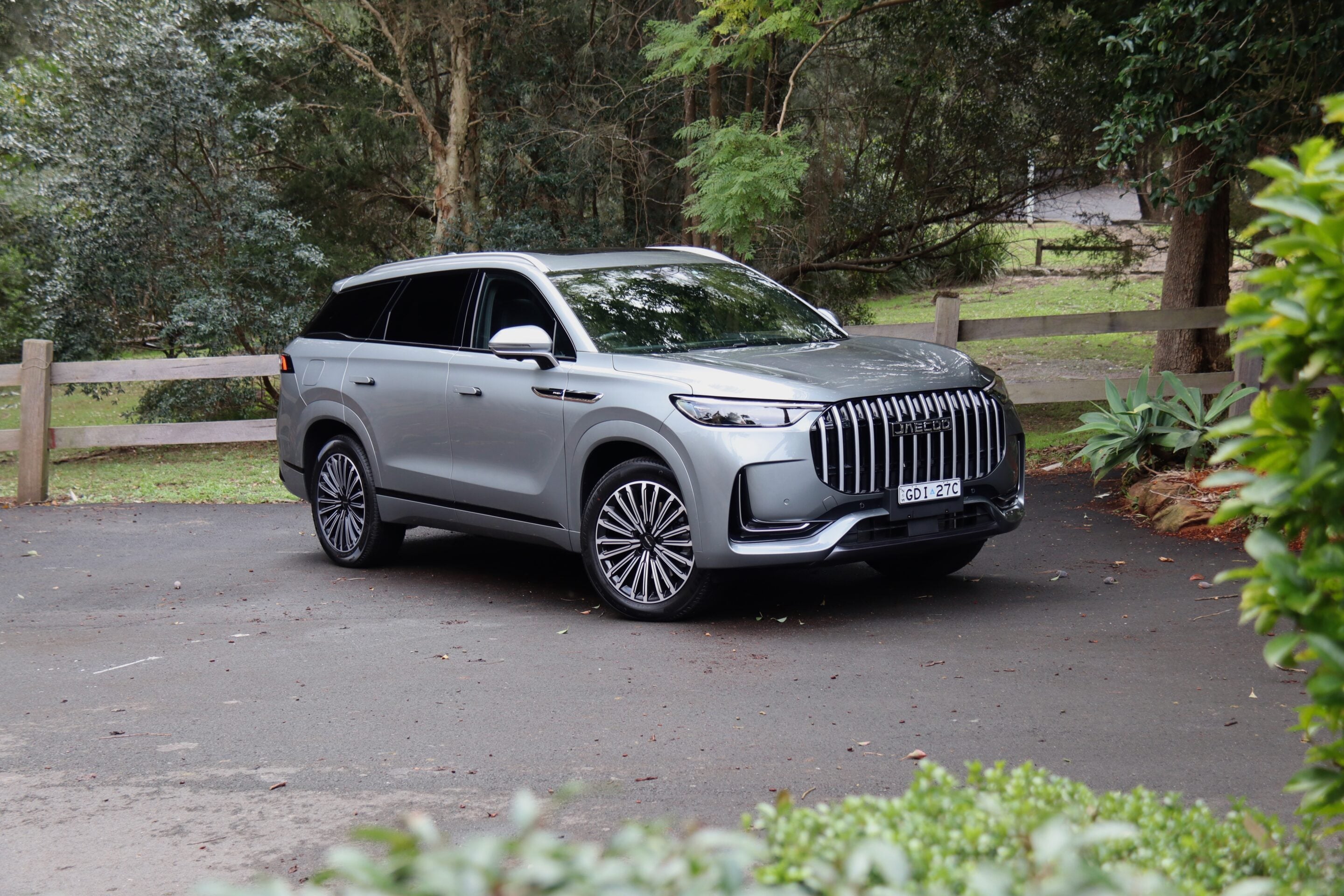 Silver SUV parked on a paved lot with a wooden fence and trees in the background, reflecting the surroundings on its windows and hood.