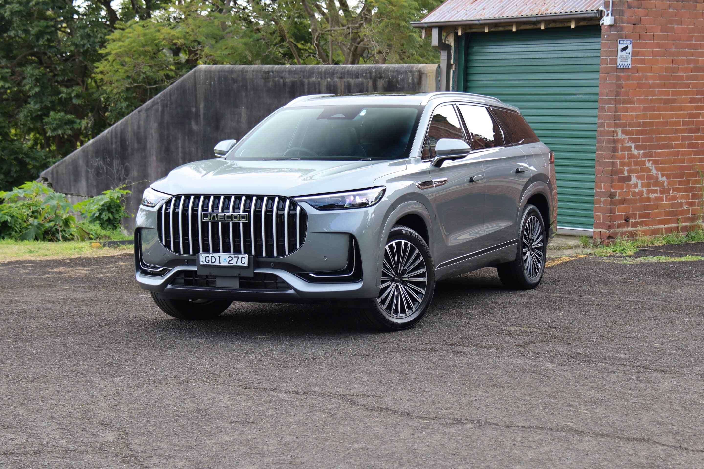 Silver SUV parked in a driveway beside a brick building with a green rolled door and trees in the background.