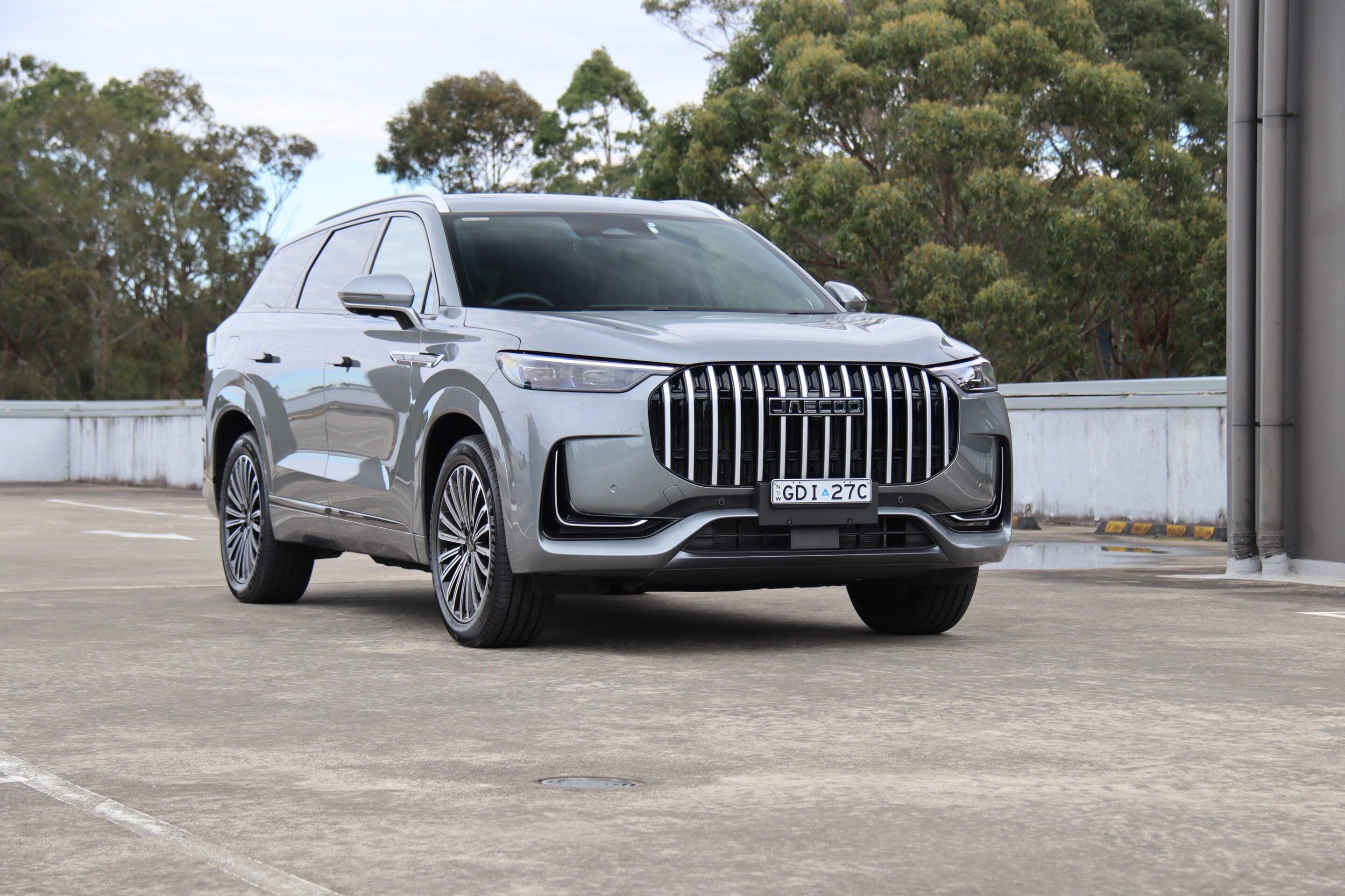 Front three-quarter view of a silver SUV with a bold vertical grille parked in an outdoor lot, trees in the background.