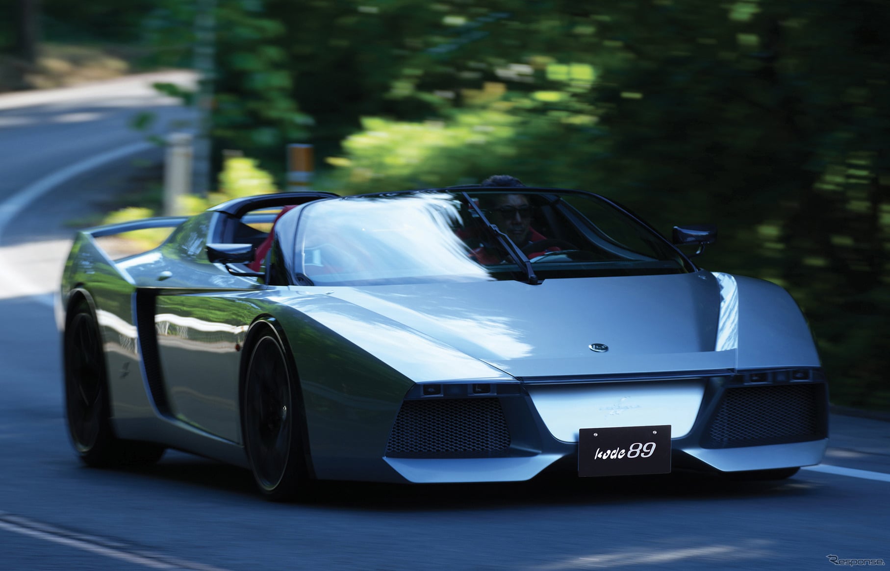 Silver convertible sports car in motion on a winding road, with the driver visible through the windshield.