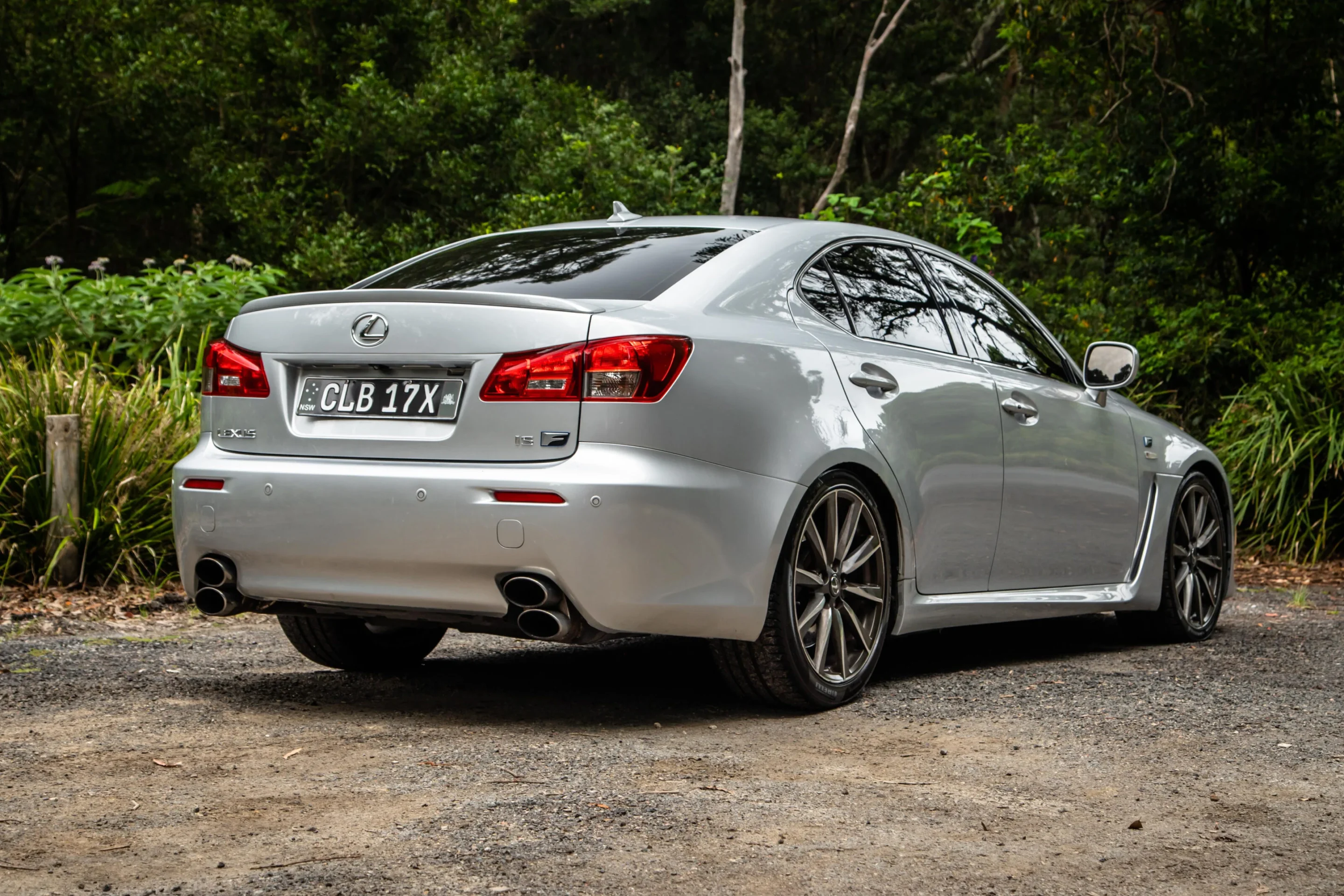 Rear three-quarter view of a silver Lexus sedan parked on a gravel driveway, with green shrubs and trees in the background and dual exhausts visible at the rear.