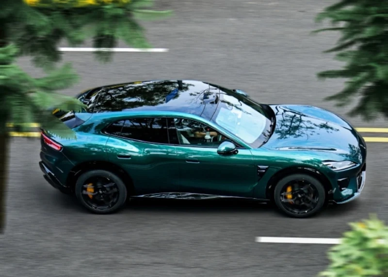 Green sporty SUV car in motion on a city street, reflections on the hood, with blurred trees and road markings suggesting speed.
