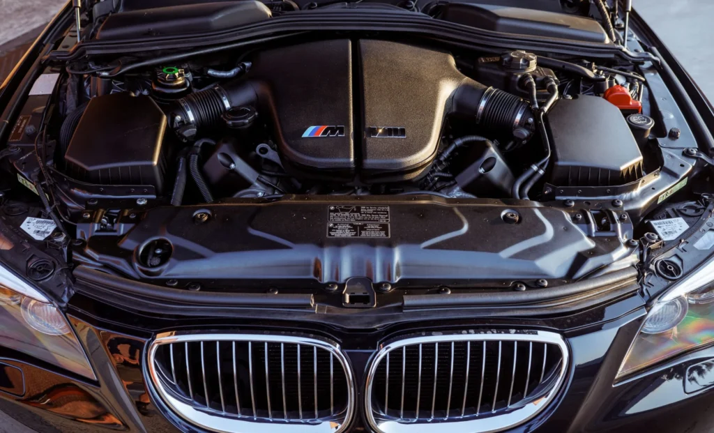 Open front view of a BMW engine bay with M logo on the intake cover, showing hoses and air boxes surrounding the engine core.