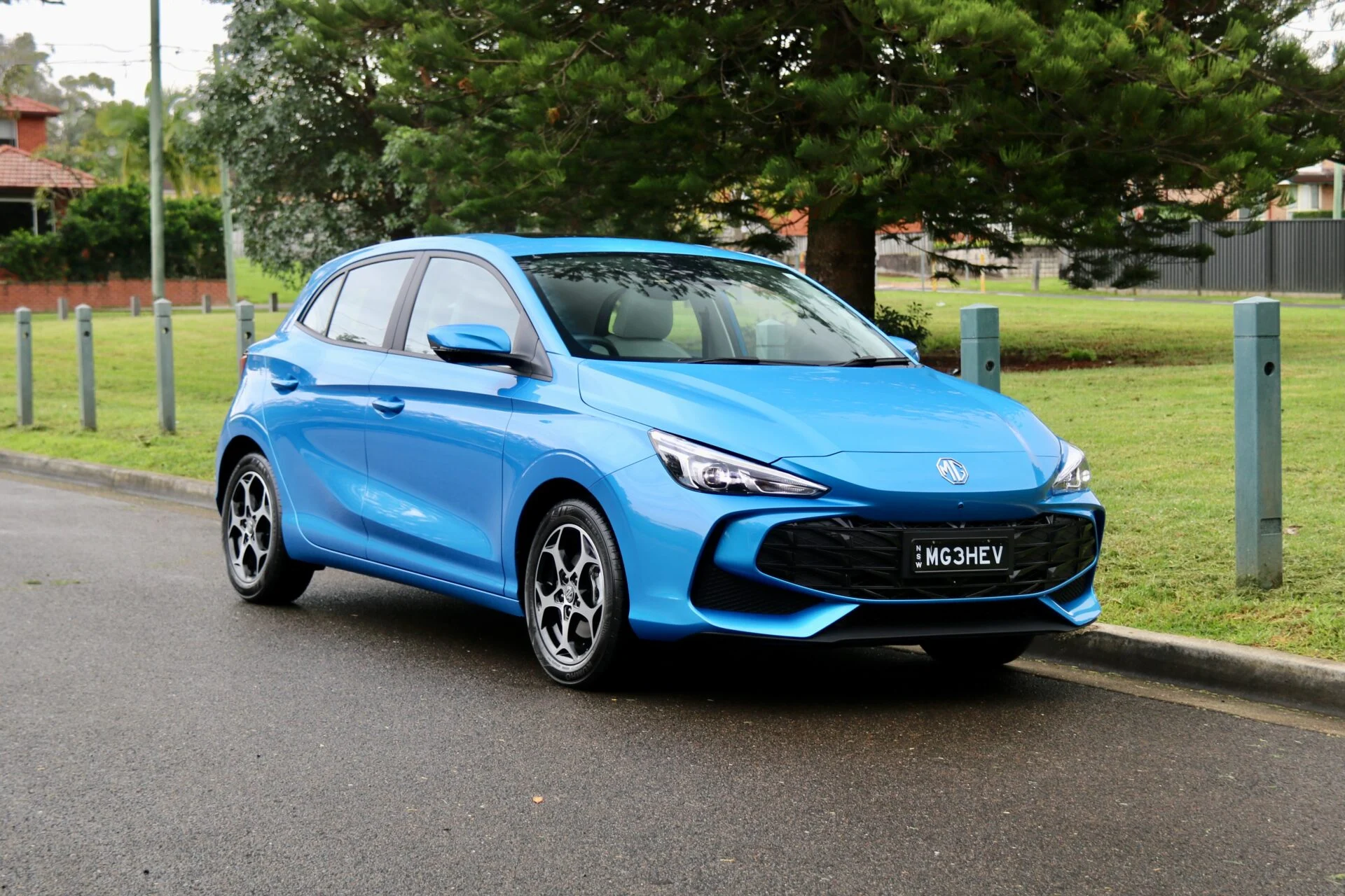 Front three-quarter view of a blue MG hatchback parked along a curb with a grassy area and trees in the background, license plate MG3HEV.