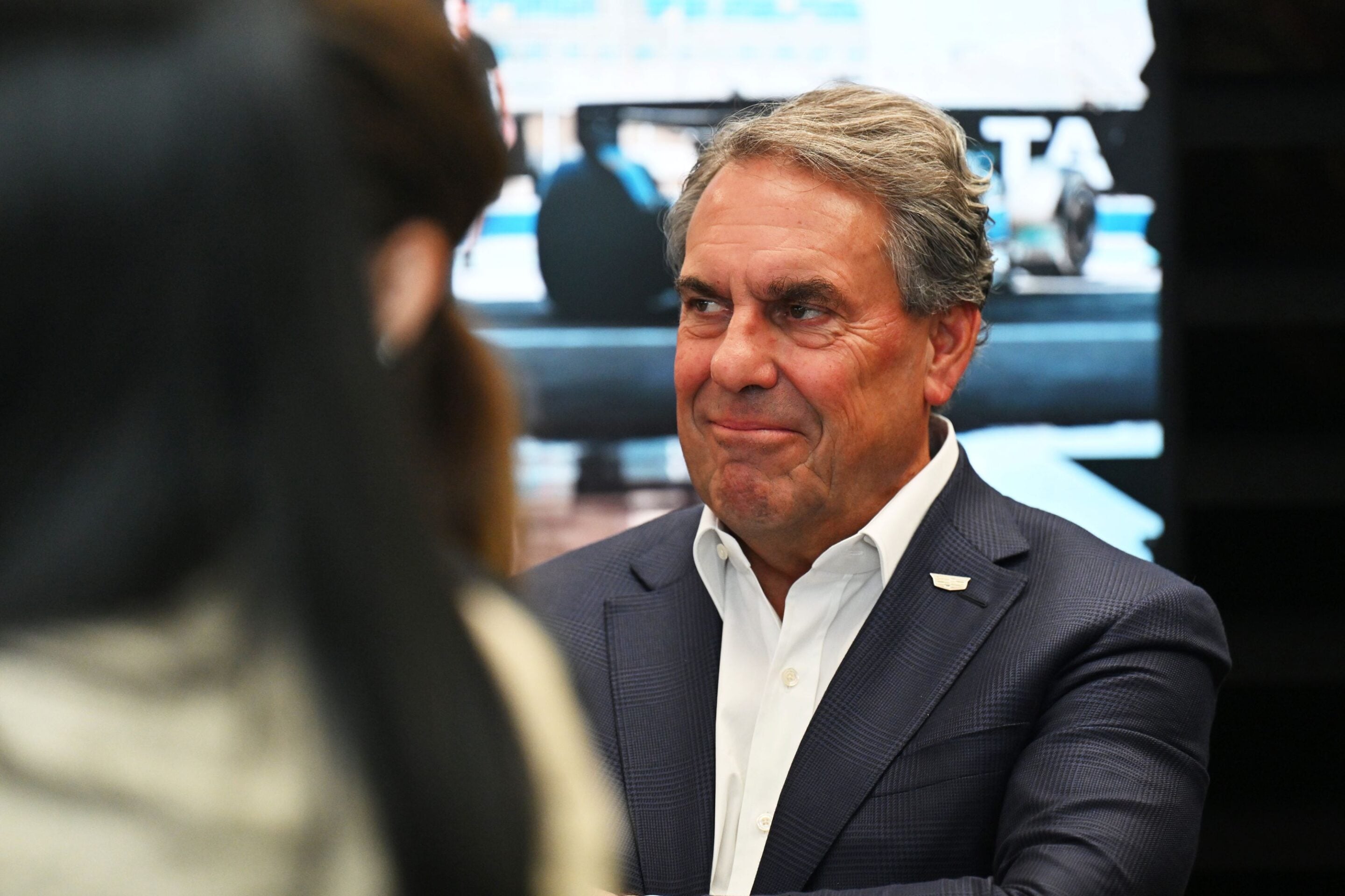 Middle-aged man with gray hair in a dark suit and white shirt, seated and engaged in conversation at a formal event.