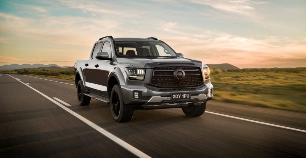 Gray pickup truck driving along an open highway at sunset, desert hills in the background, front three-quarter view