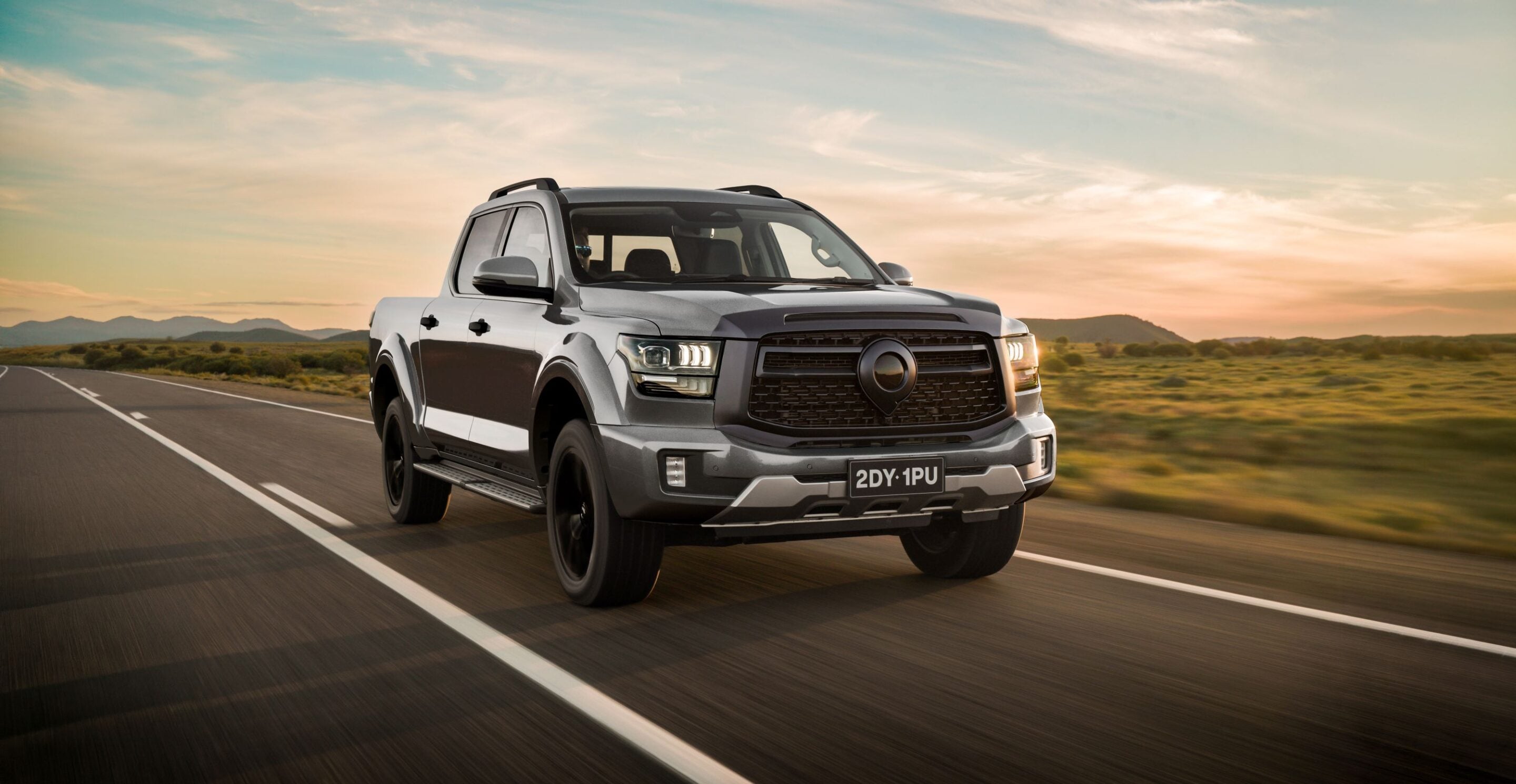 Gray pickup truck driving along an open highway at sunset, desert hills in the background, front three-quarter view