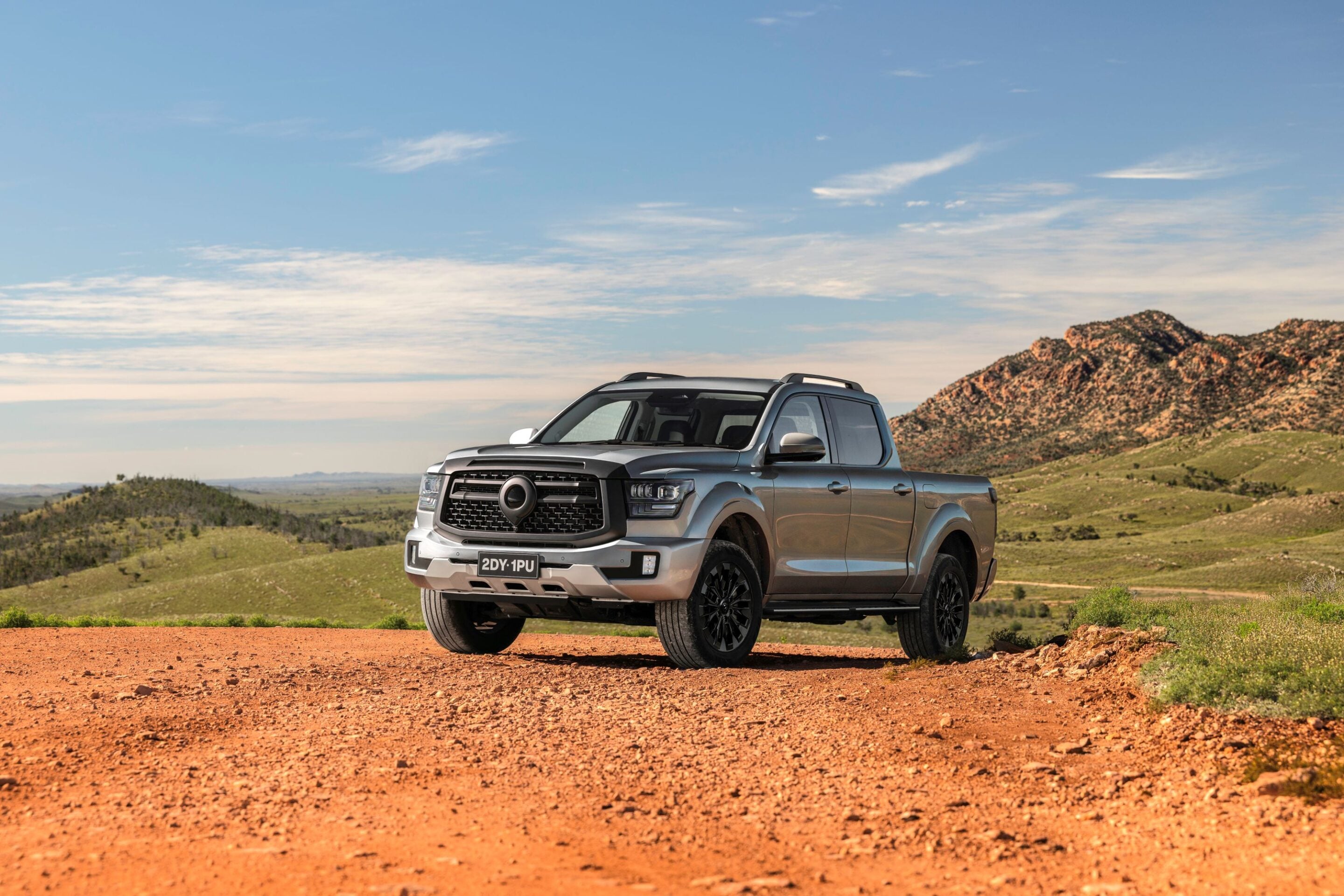 Front three-quarter view of a silver pickup truck parked on a dusty red dirt road with rolling hills and a blue sky behind it.