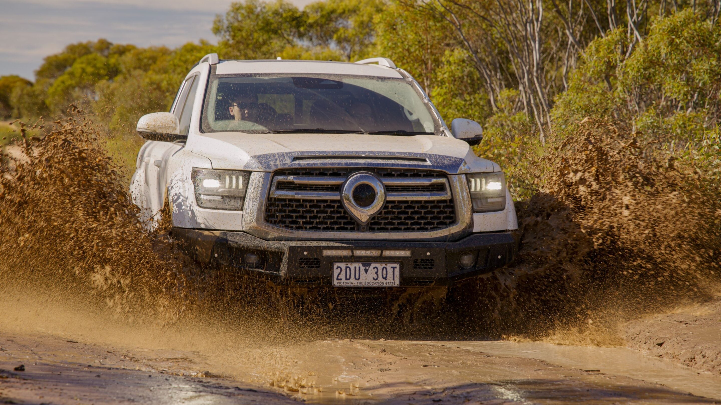 White SUV splashes through a muddy puddle on a dirt road with trees in the background.