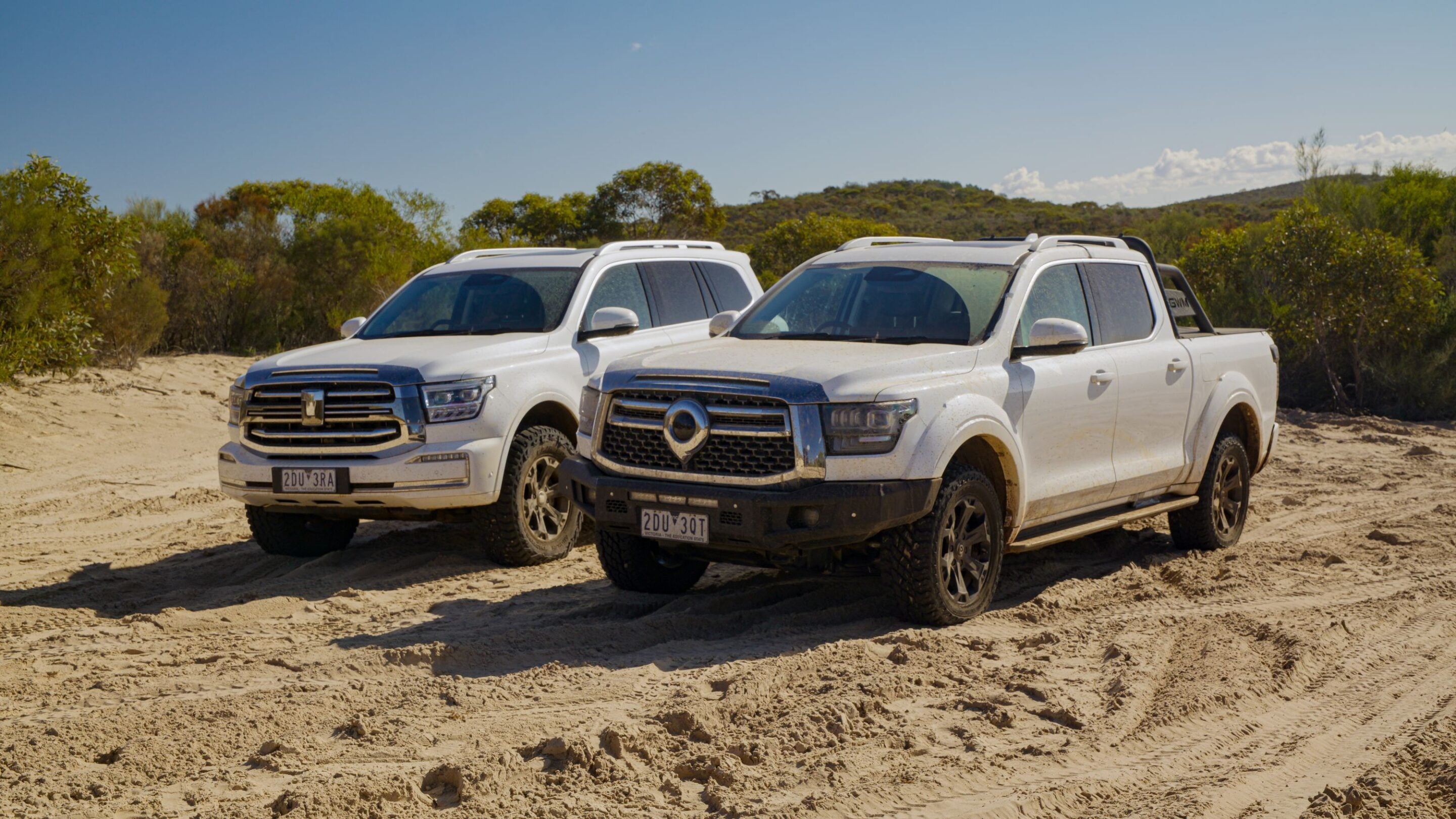 Two white pickup trucks parked side by side on a sandy desert trail with shrubs and a blue sky above.