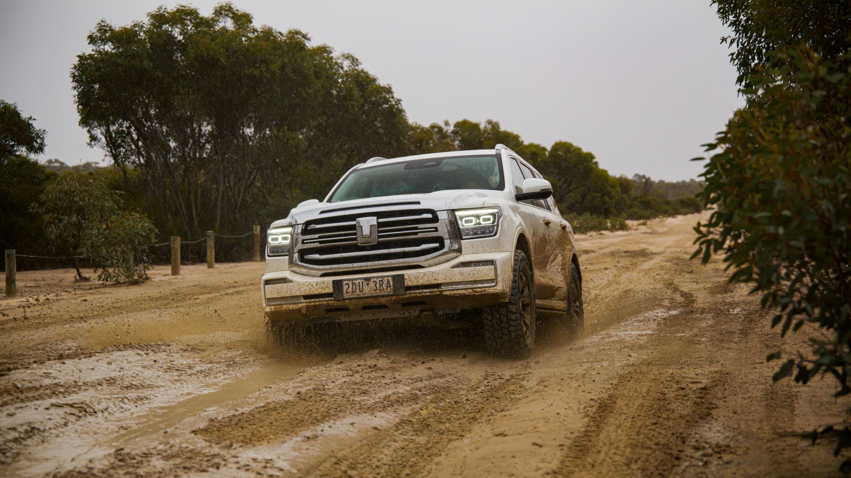 White pickup truck driving through a muddy dirt road, mud splashing around the wheels in a rural, tree-lined landscape.