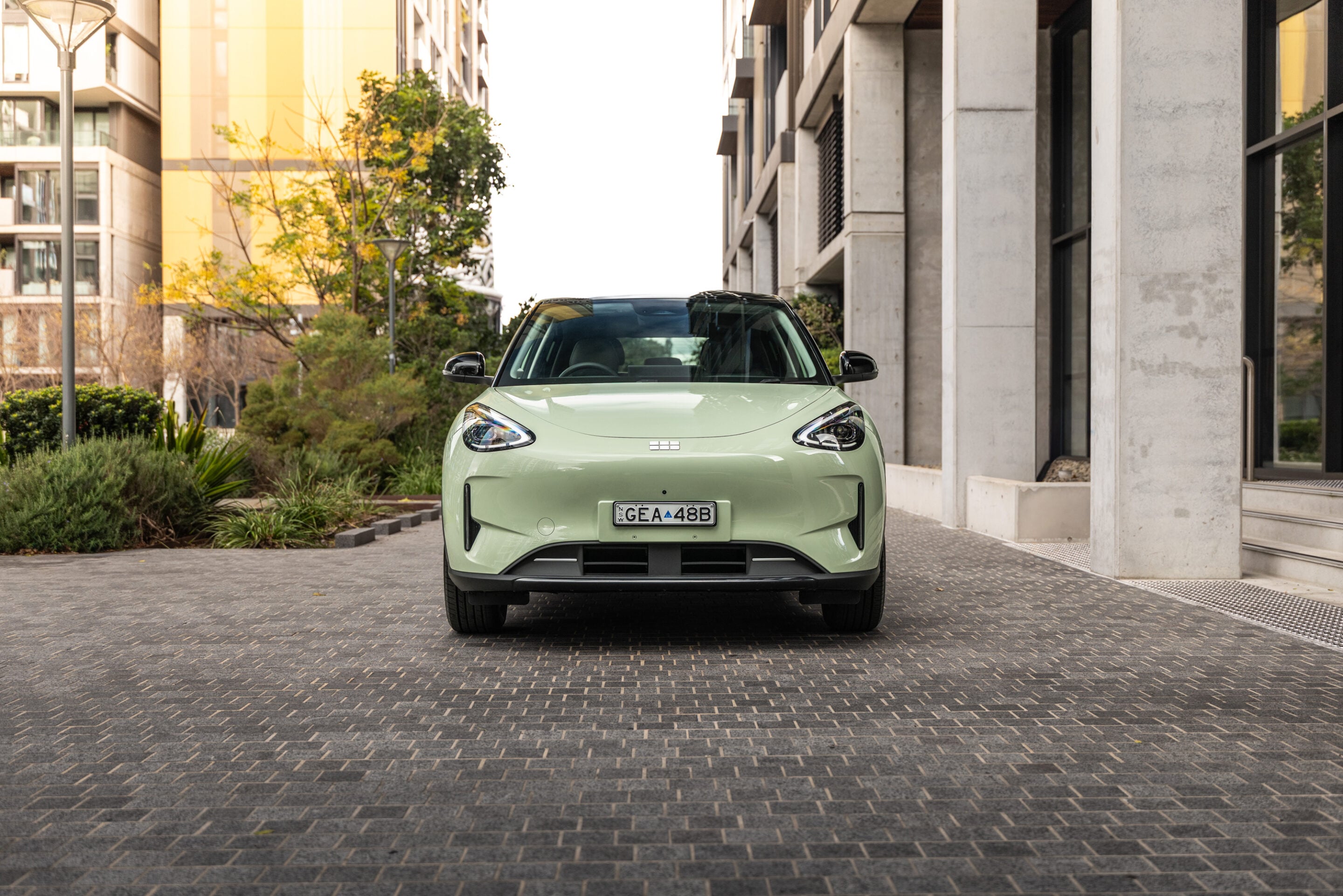 Front view of a light green compact car parked on a brick-paved driveway between modern buildings.