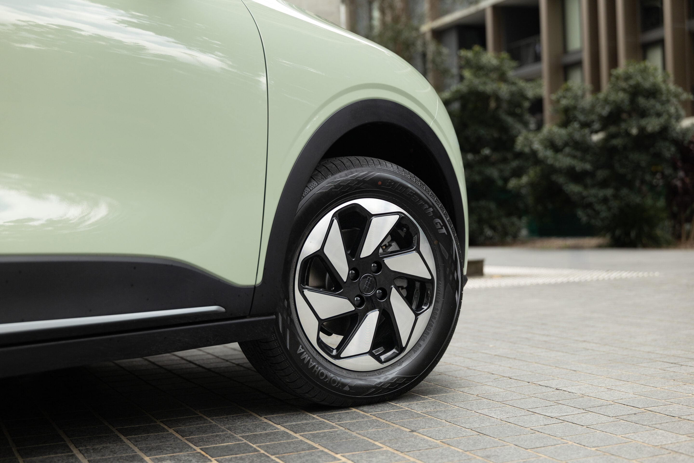 Close-up of a pale green car's front wheel and fender, showing the black wheel arch trim and alloy wheel on a brick-paved surface.