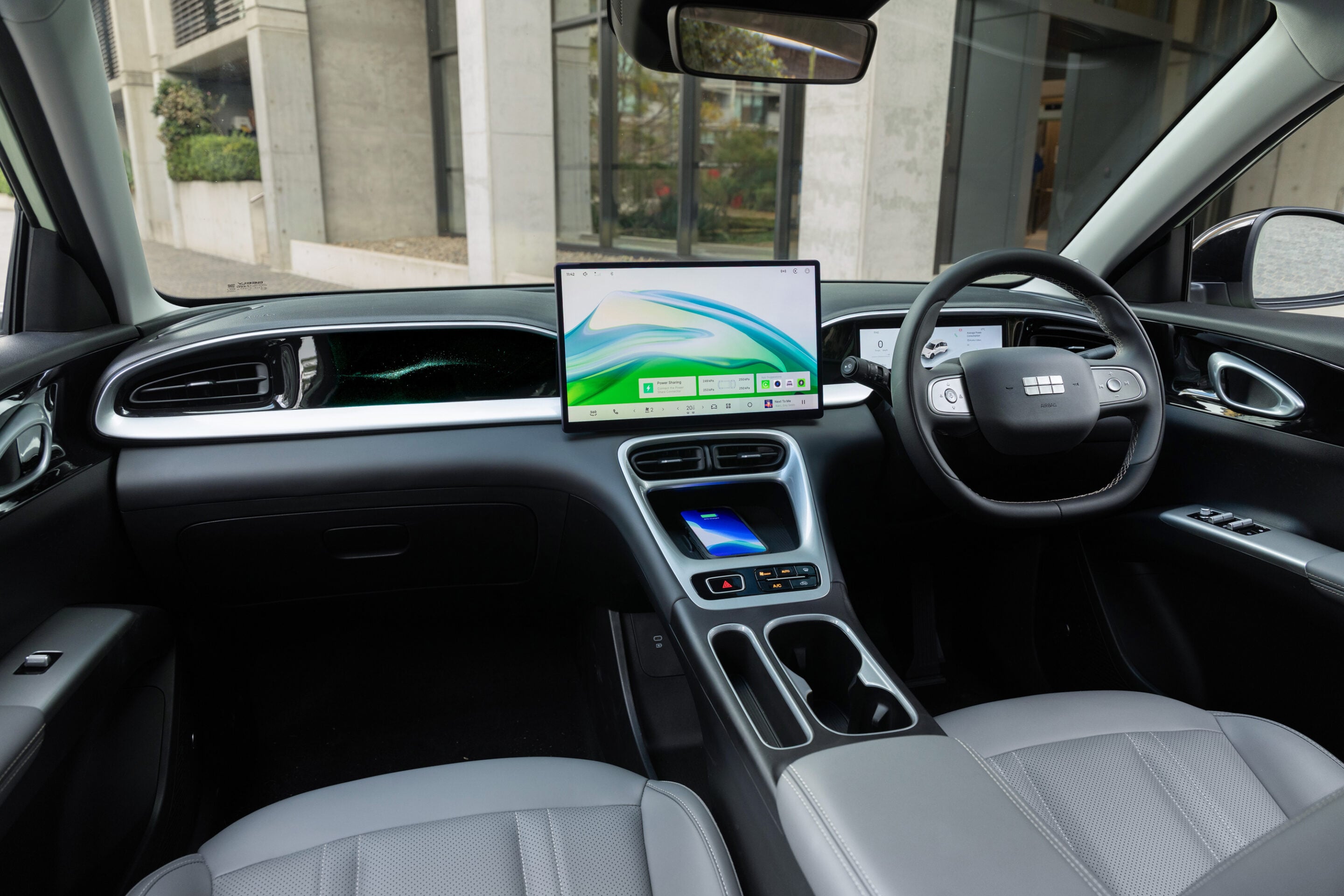 Modern car cabin with a large center touchscreen, steering wheel on the right, and gray leather seats.