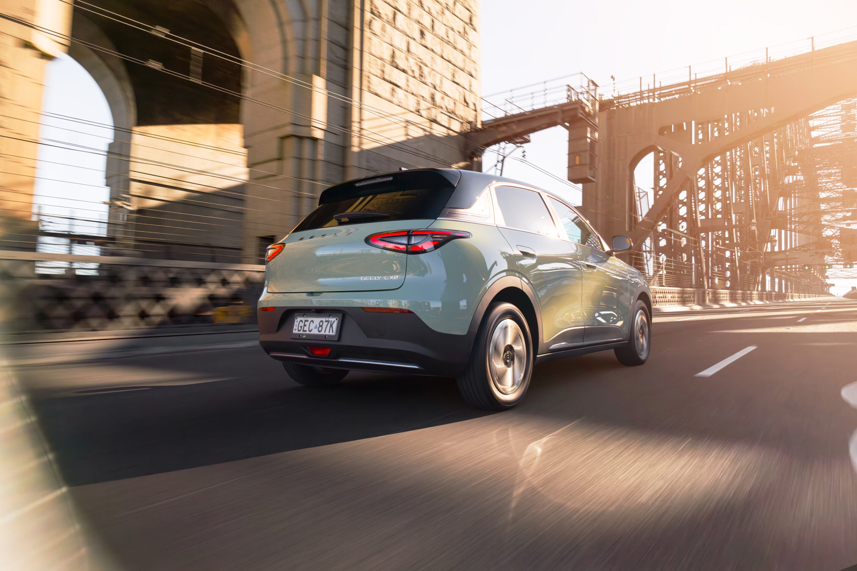 Rear of a light blue SUV driving on a bridge at golden hour, with steel arches and overhead cables in the background.