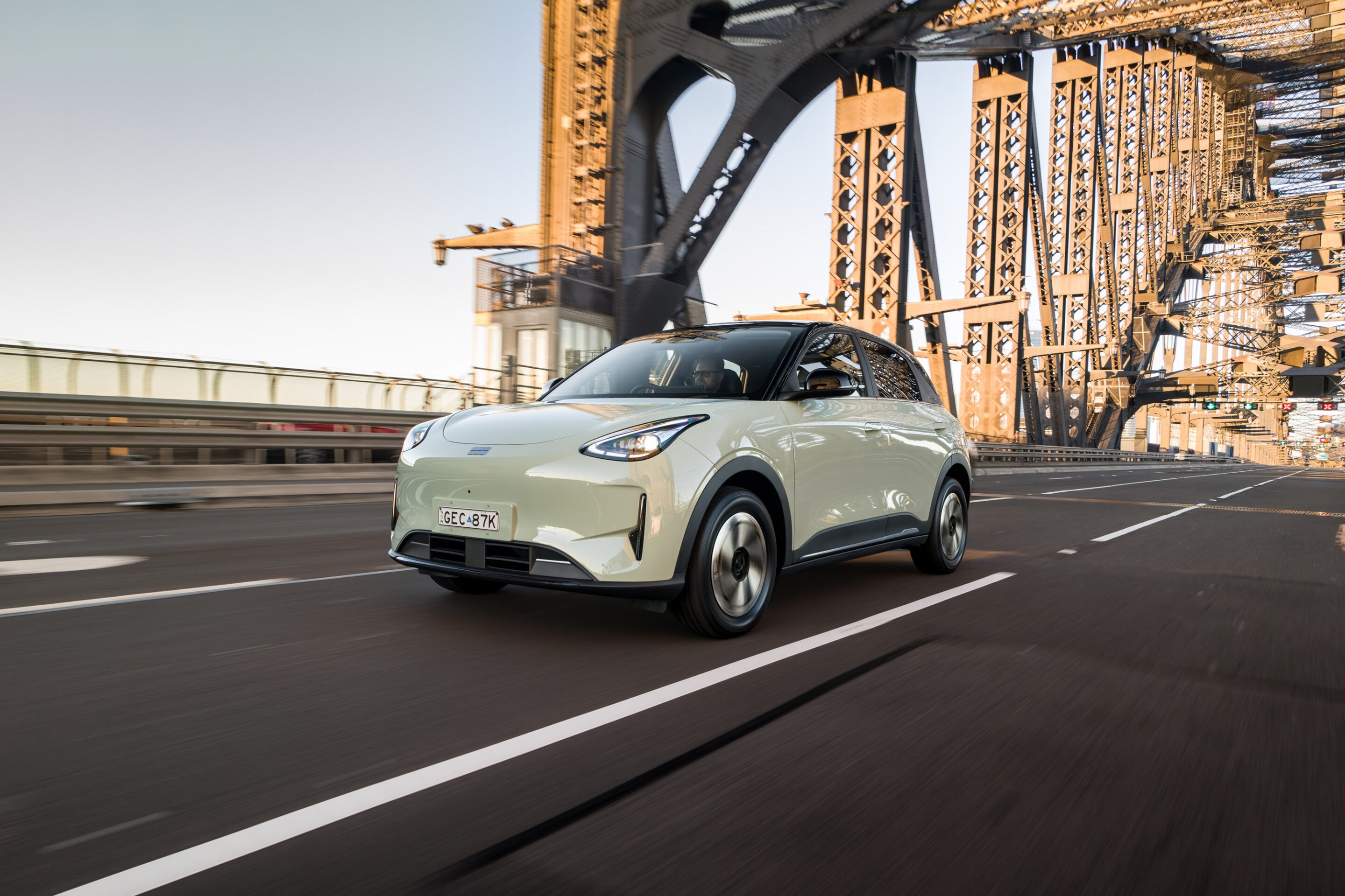 White compact electric car driving on a wide steel-truss bridge, motion blur on the road and warm daylight illuminating the structure.