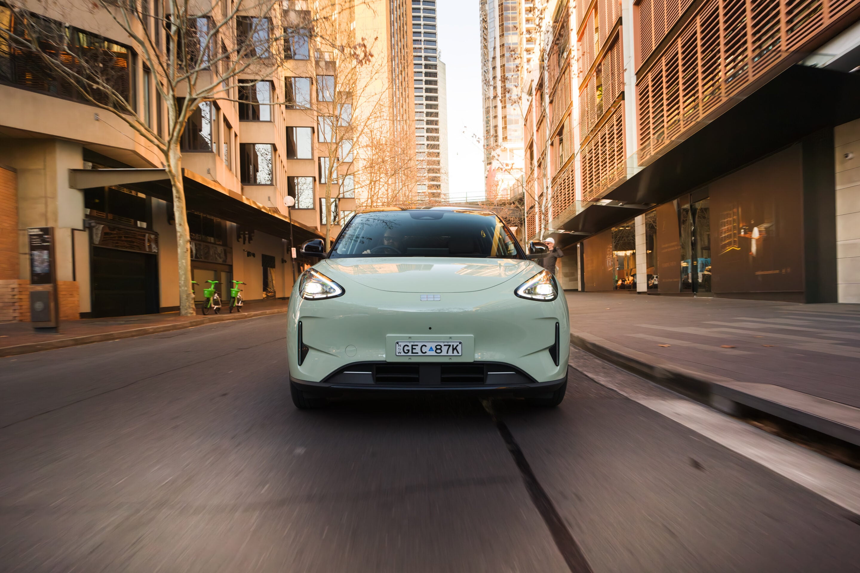 Mint-green electric car drives toward camera on a narrow city street, tall buildings towering on both sides with motion blur behind it.