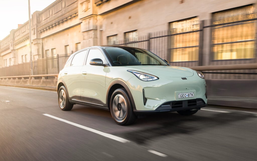 Mint-green crossover SUV driving quickly along a city street with blurred beige buildings in the background at sunset.