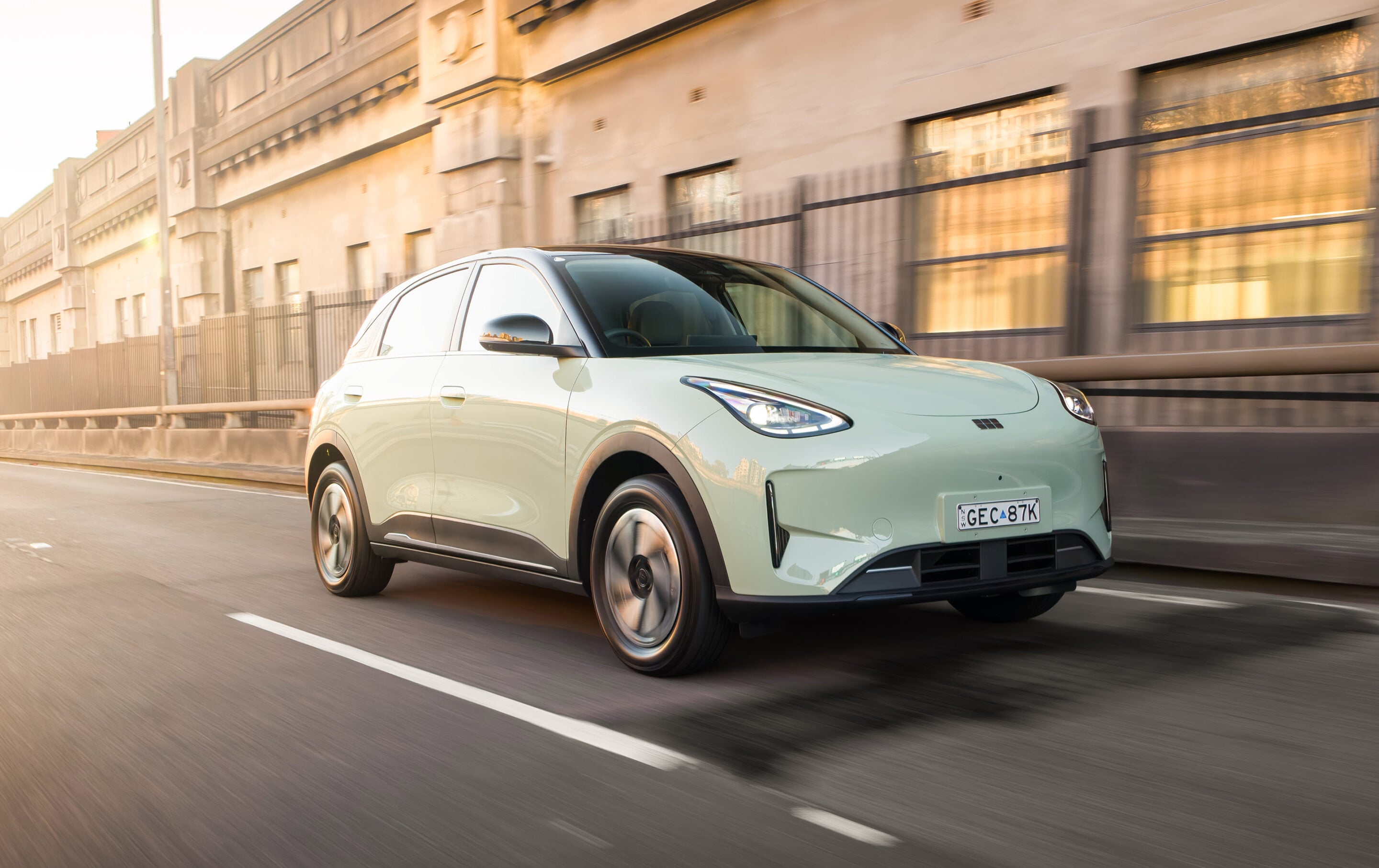 Mint-green crossover SUV driving quickly along a city street with blurred beige buildings in the background at sunset.