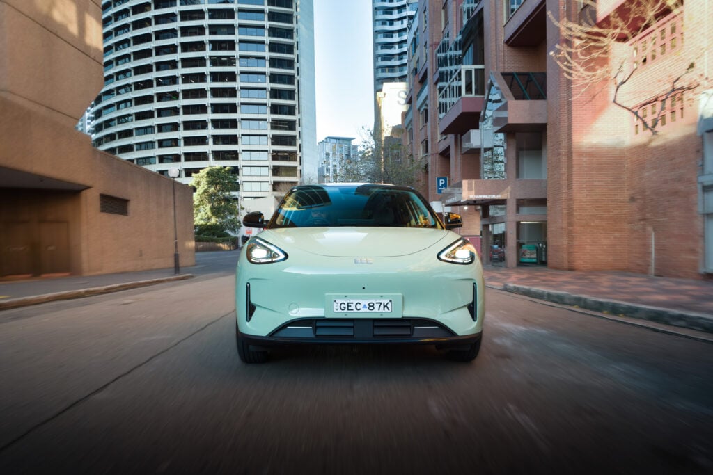 Rear view of a mint-green car driving toward the camera on a narrow urban street, tall buildings on both sides.