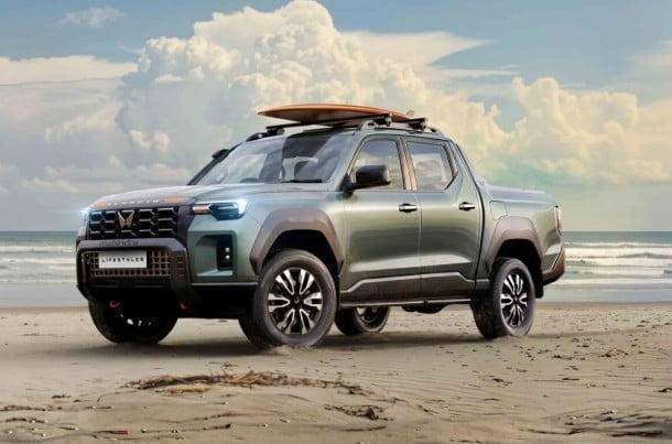 Green pickup truck with a surfboard on the roof parked on a sandy beach by the ocean.