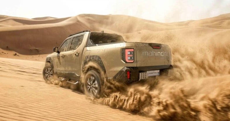 Mud-splattered Mahindra pickup charging through desert dunes, kicking up a spray of sand behind it.